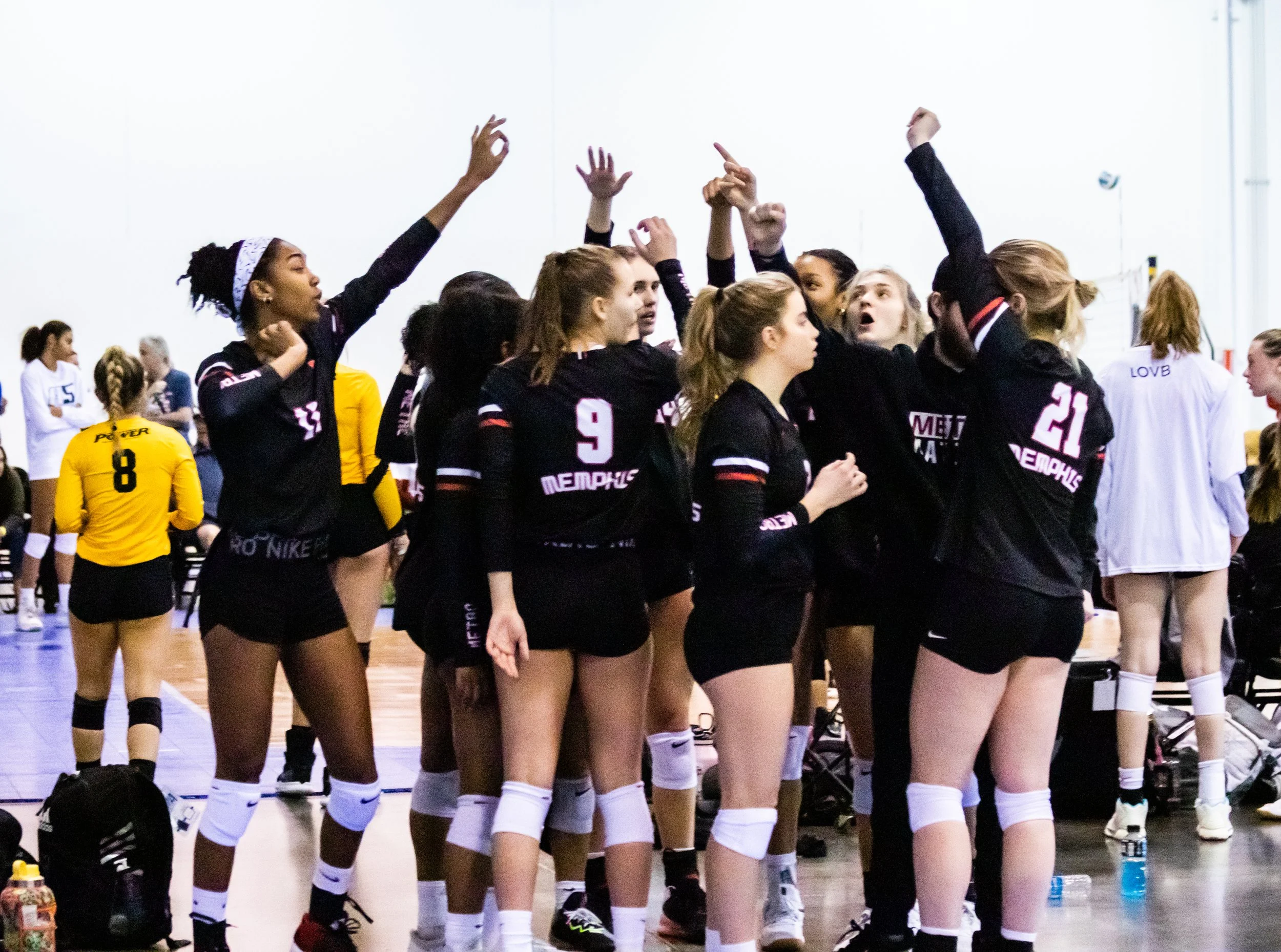 A group of female volleyball players in black uniforms celebrating together in an indoor gym.