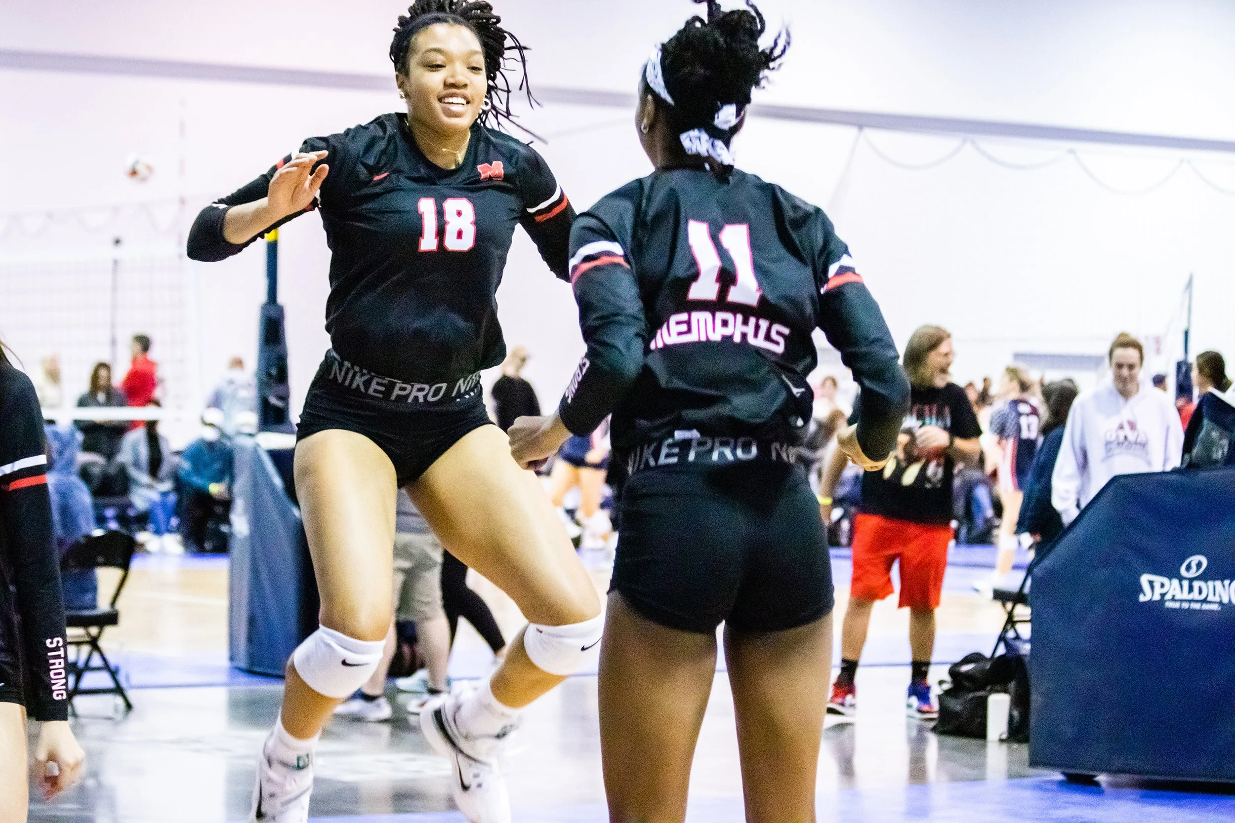 Two female volleyball players in black uniforms with pink and white accents, celebrating on an indoor court. One is jumping with a smile, the other stands with her back to the camera. The court and spectators are visible in the background.