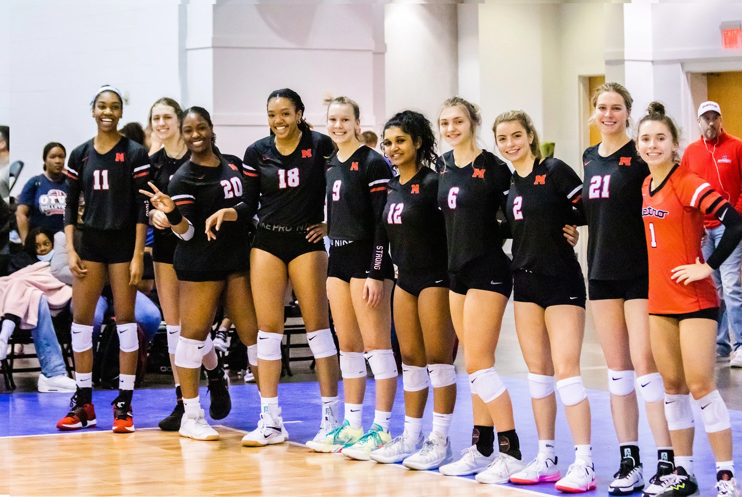 A group of nine female volleyball players standing in a line on an indoor court, wearing black and orange uniforms with knee pads, smiling for a team photo.