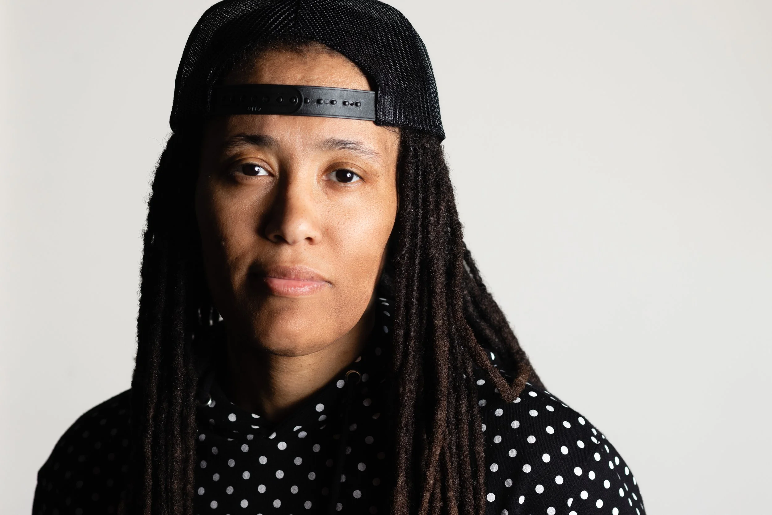 Close-up portrait of a woman with dreadlocks wearing a black and white polka dot shirt and a black baseball cap backwards against a plain light background.