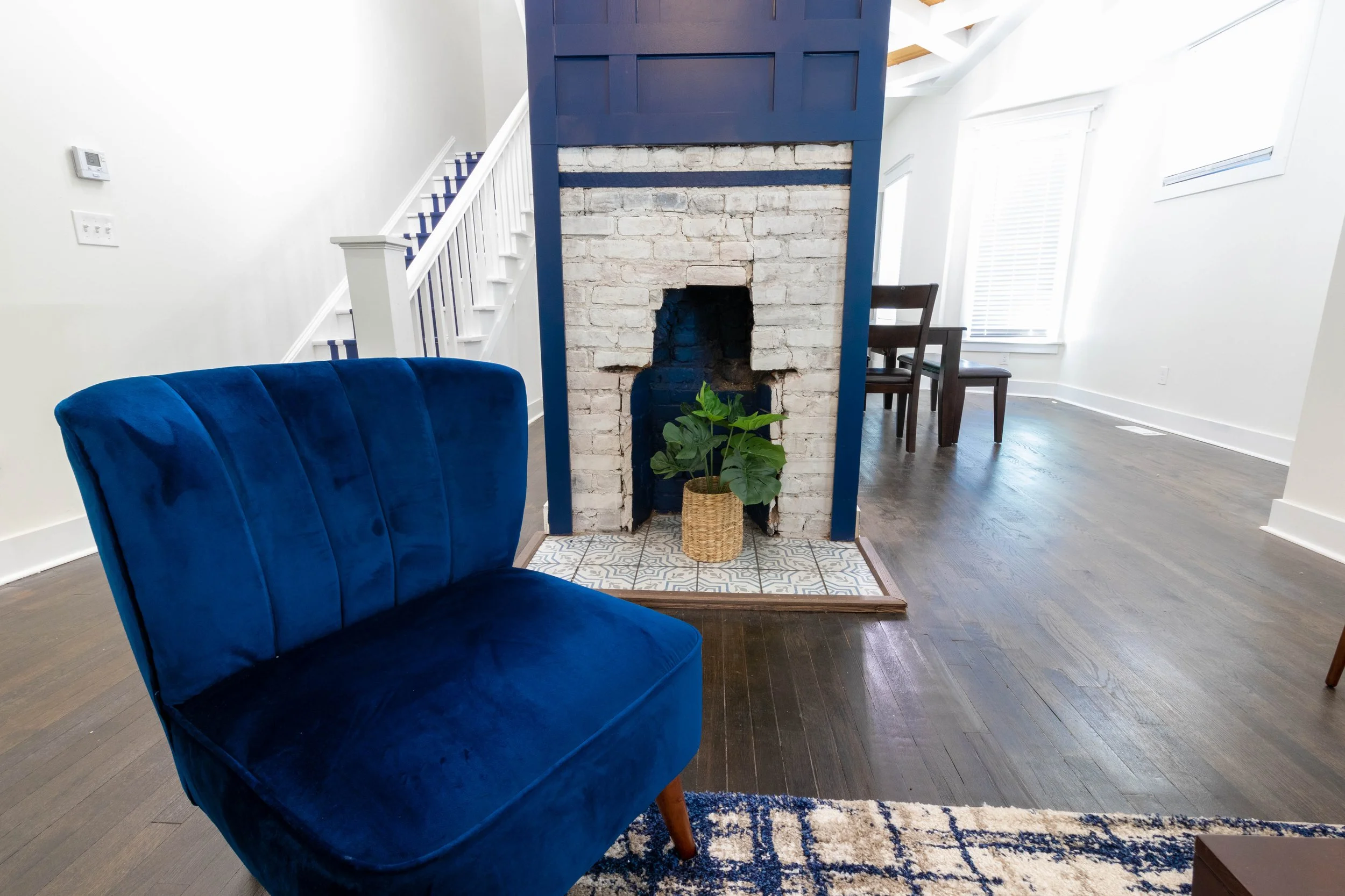 Living room with white walls, a blue velvet armchair, hardwood floors, a brick fireplace with blue paneling, a potted plant, and a dining area in the background.