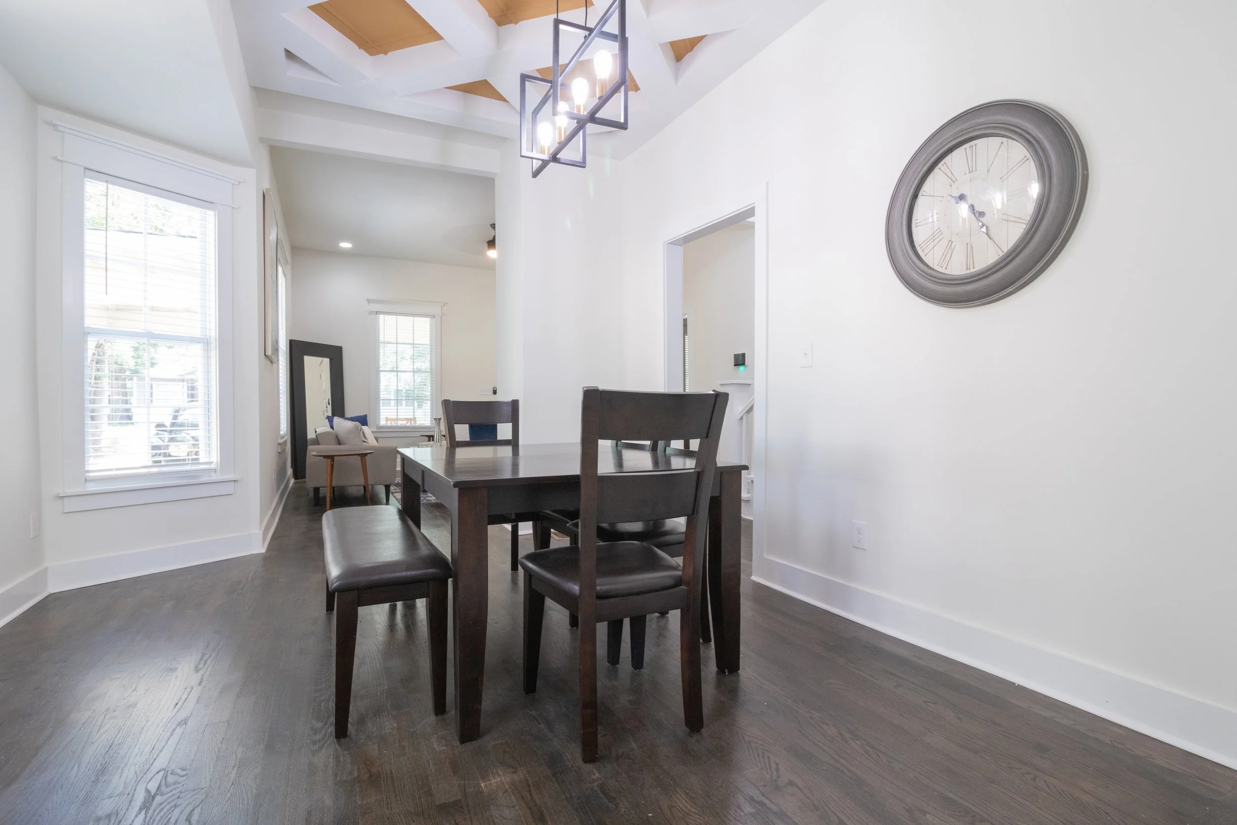 Dining area with dark wood table and chairs, white walls, large wall clock, hardwood flooring, windows with white trim, and a modern chandelier.