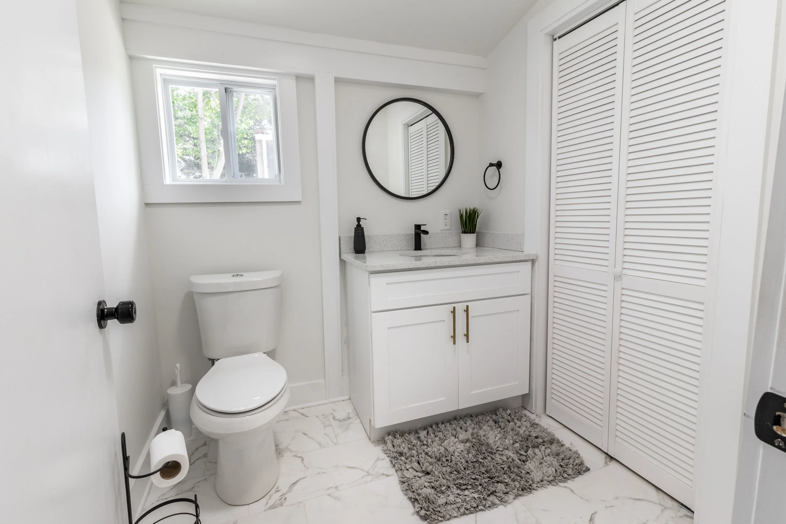 A modern white bathroom with a toilet, a black soap dispenser on a white vanity with gold handles, an oval mirror, a small window, a gray shaggy rug, and a closet with louvered sliding doors.