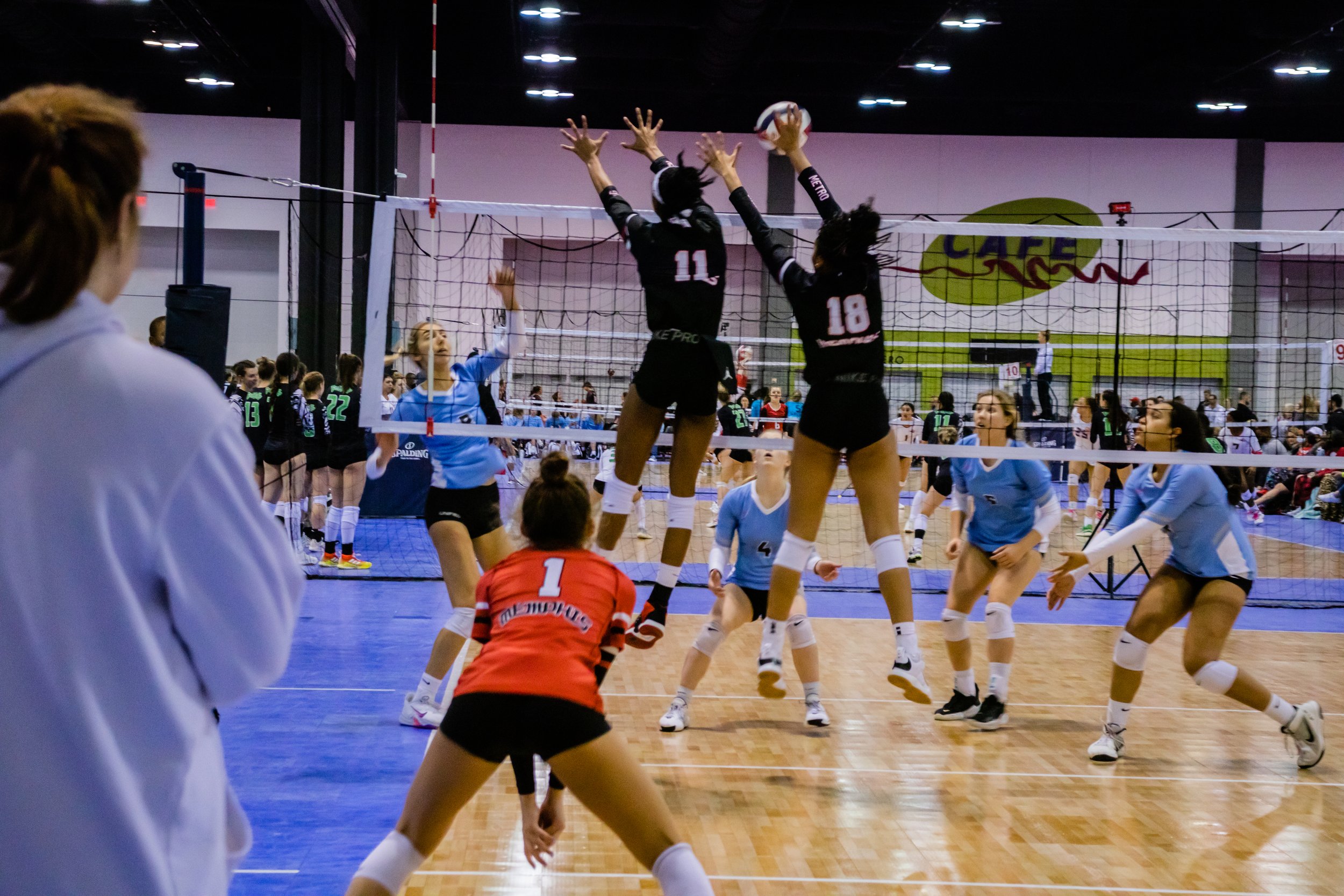 A volleyball game in progress with players jumping to block and spike the ball at the net, while others are preparing to receive or defend, inside an indoor gymnasium.