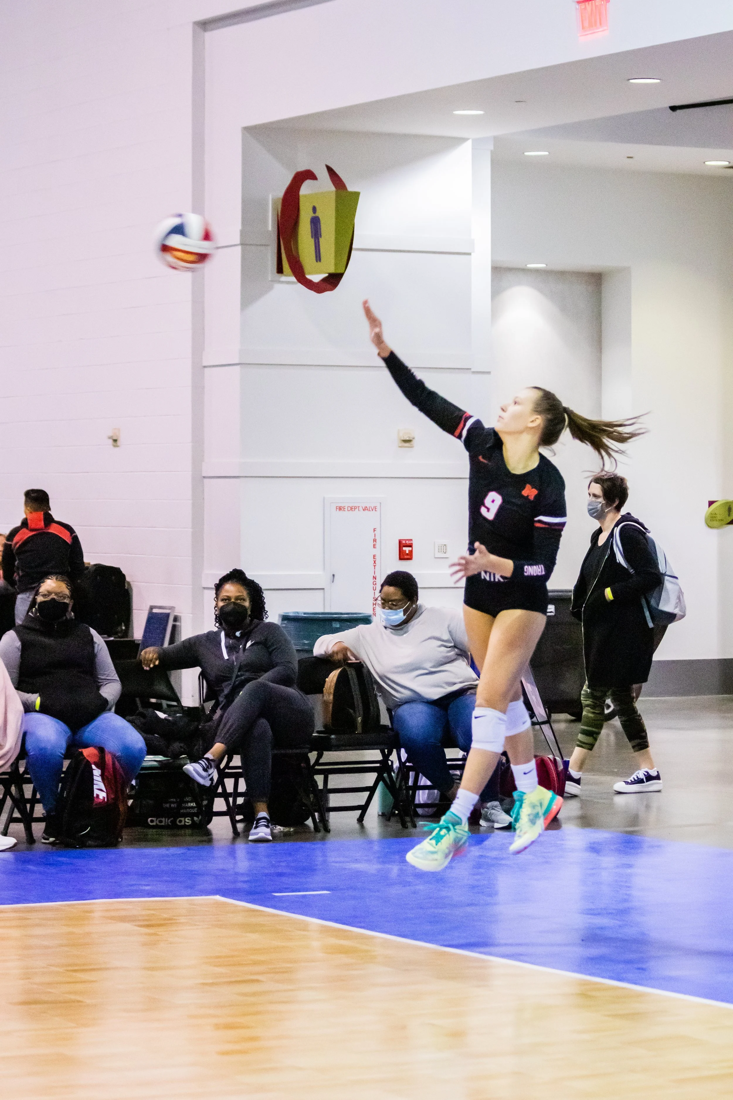 A female volleyball player wearing a black jersey with the number 9, white knee pads, and teal sneakers is jumping to hit a volleyball in an indoor court. Several spectators, all wearing masks, are sitting and standing nearby watching her play.