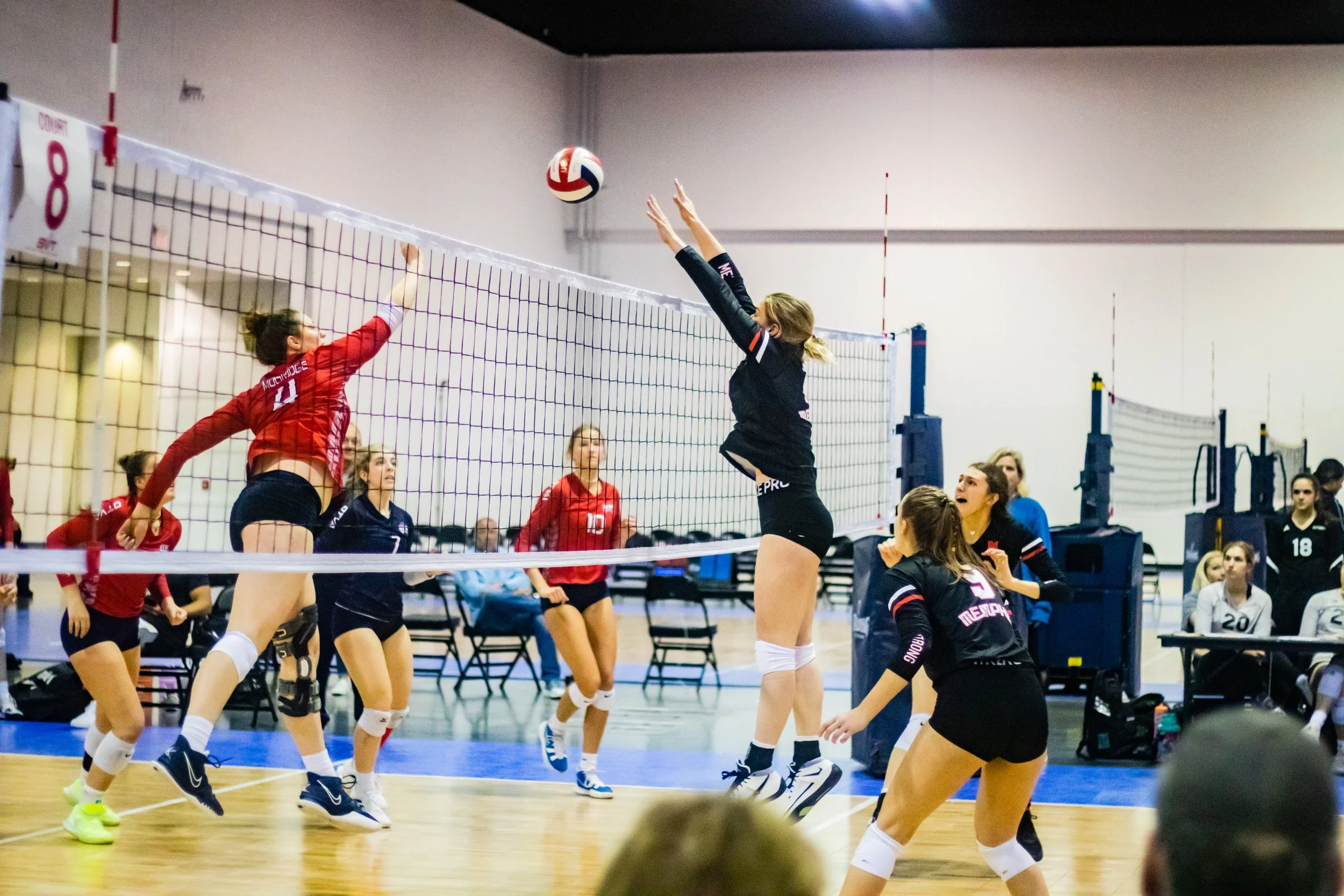 A volleyball match with players from two teams, one in red and one in black, actively blocking and spiking the ball at the net inside an indoor gym.