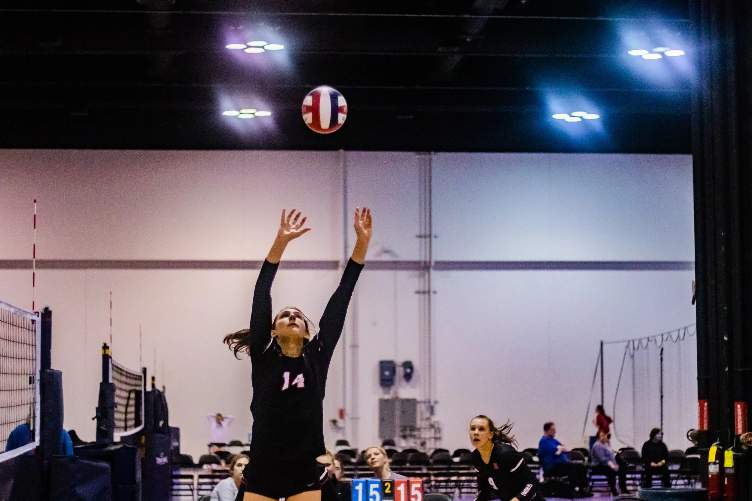A female volleyball player in a black uniform with the number 14 hits a volleyball during a match in an indoor gym, with other players and spectators in the background.