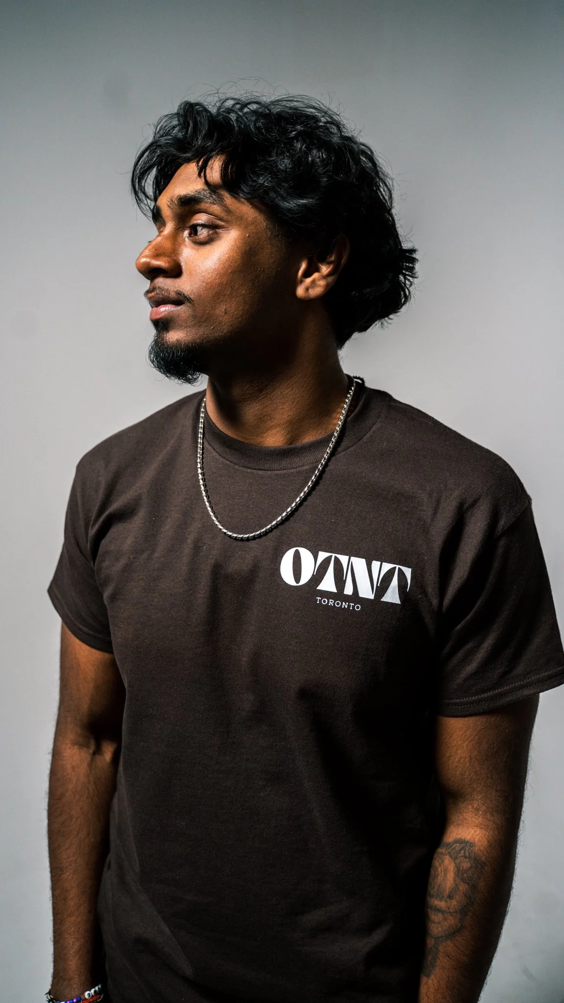 A young man with dark curly hair and a goatee wearing a black t-shirt with the word 'OTNT' and 'Toronto' printed on it, accessorized with a silver chain necklace and bracelets, standing against a plain grey background.
