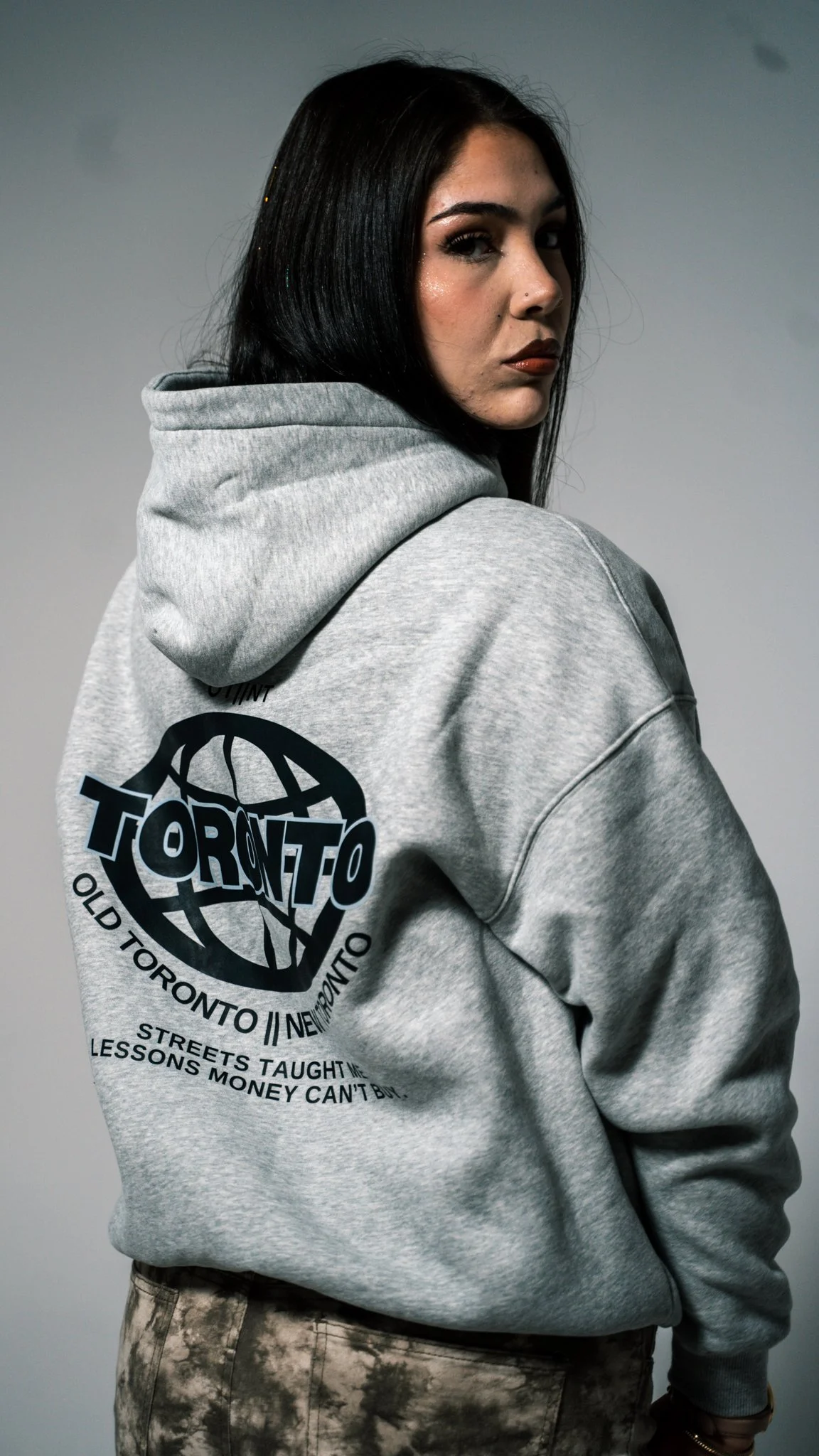 A woman with long dark hair wearing a gray Toronto hoodie and camouflage pants, looking over her shoulder against a plain gray background.