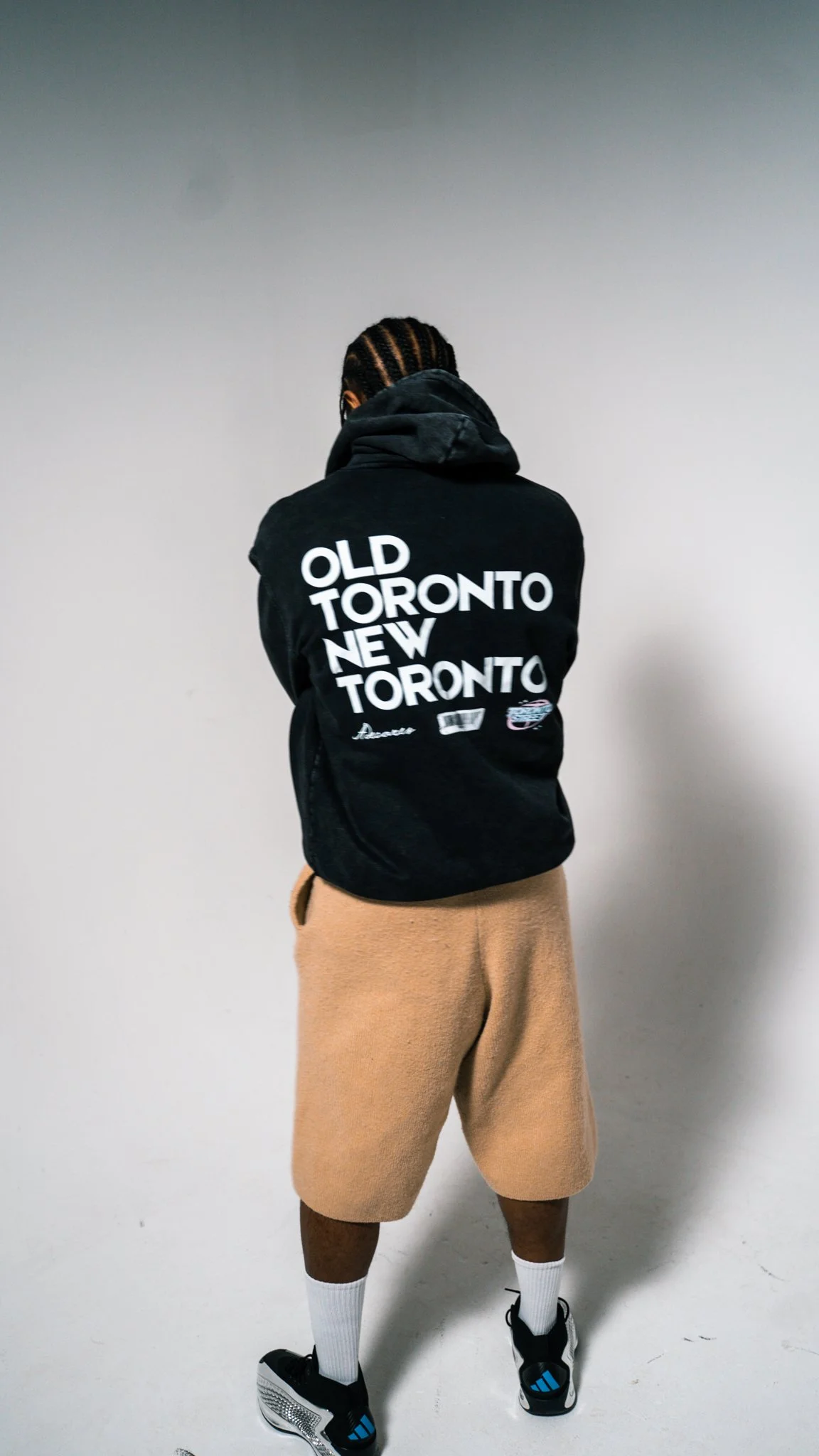 Person wearing a black hoodie with white text on the back, tan shorts, white socks, and black sneakers standing against a plain white wall.