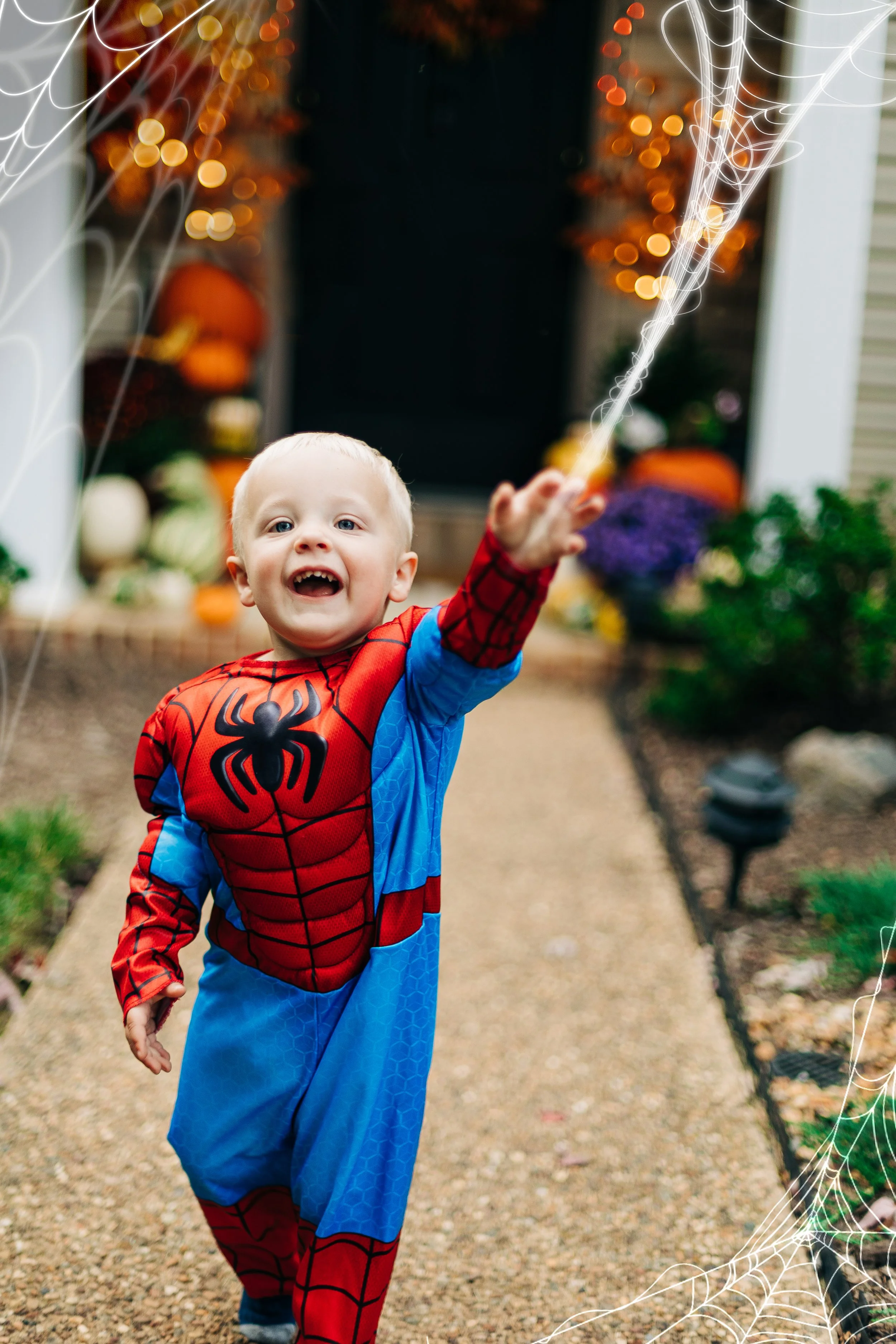 A young boy dressed in a Spider-Man costume playing with fake spider webs outside in a decorated Halloween or fall-themed yard.