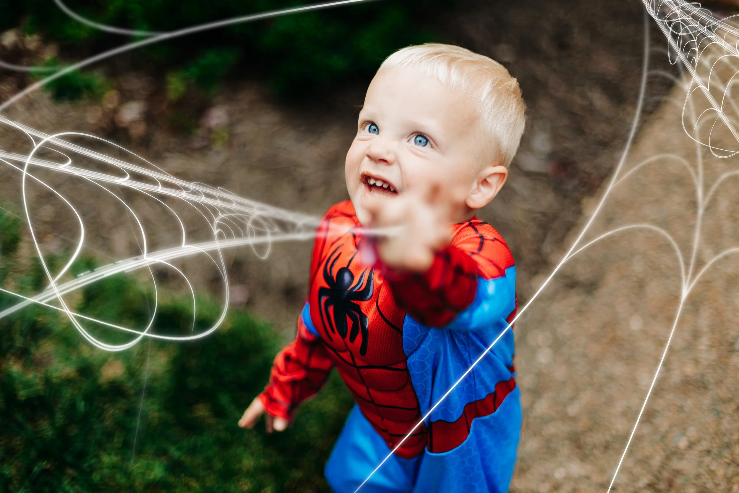Child dressed as Spider-Man reaching out and pointing, surrounded by digital web effects, outdoors on dirt and grass.
