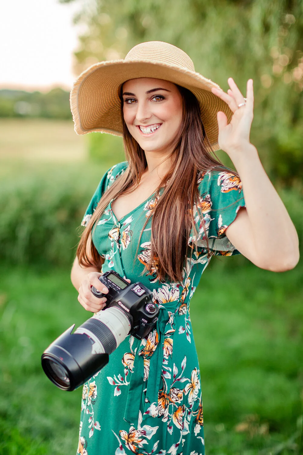 A woman in a floral dress and wide-brimmed hat smiling outdoors, holding a camera.