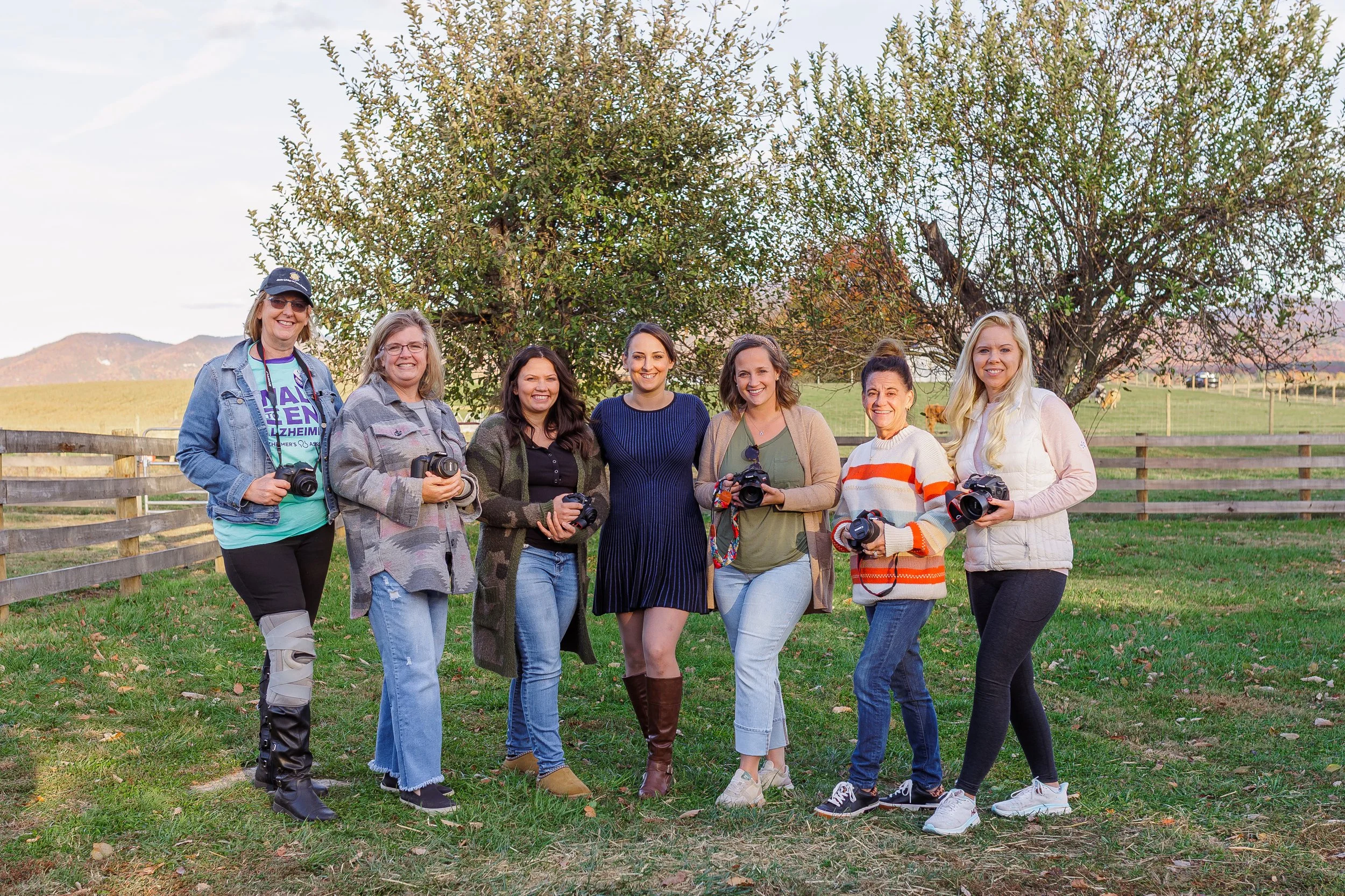 Group of seven women standing outdoors in a grassy area with a tree and mountains in the background, holding cameras and smiling.