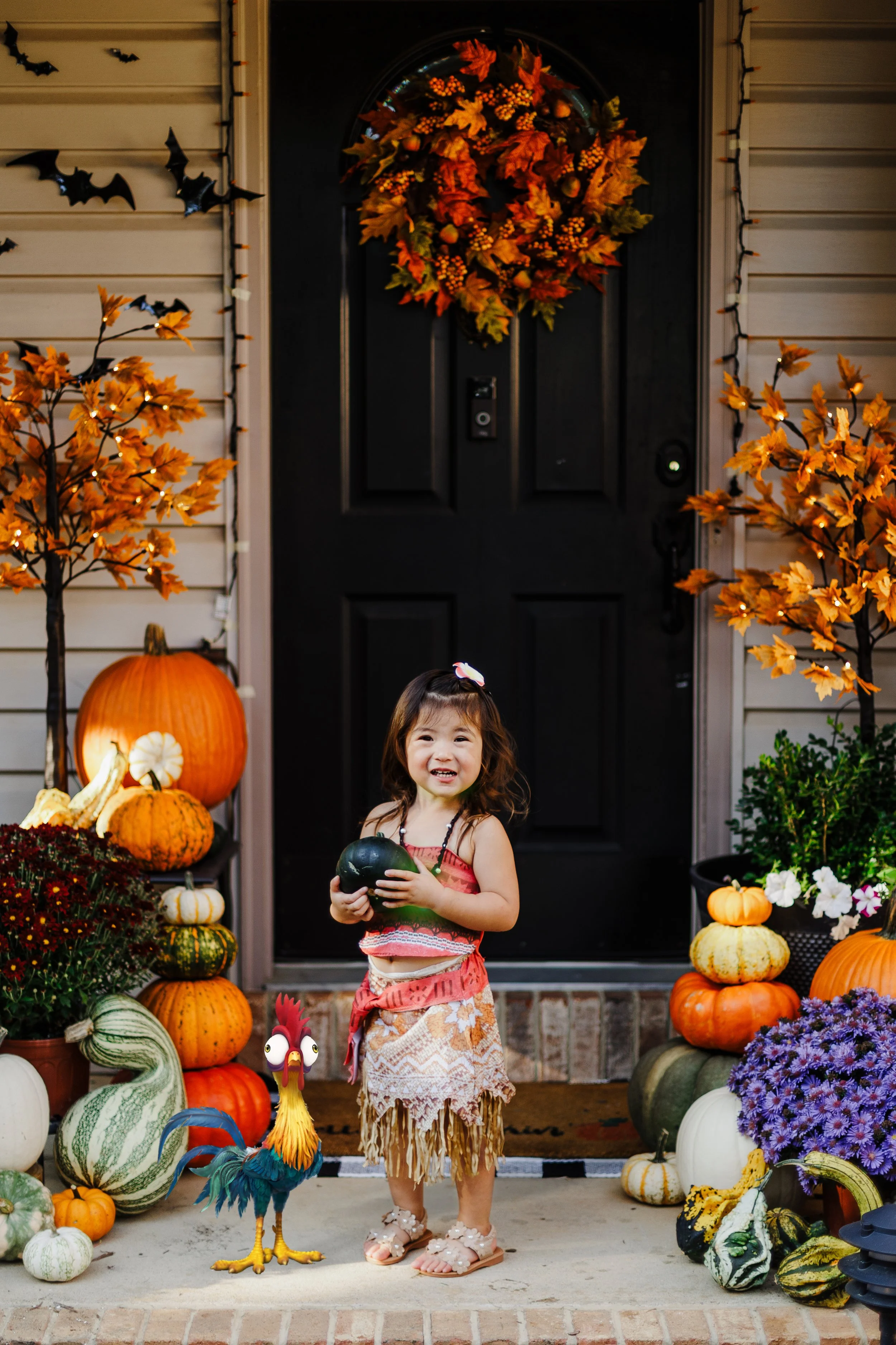 A young girl dressed in a colorful costume standing on a porch decorated with autumn-themed pumpkins, gourds, and flowers. She is holding a small pumpkin and smiling. There is a decorative rooster and autumn trees with orange leaves, a wreath on the