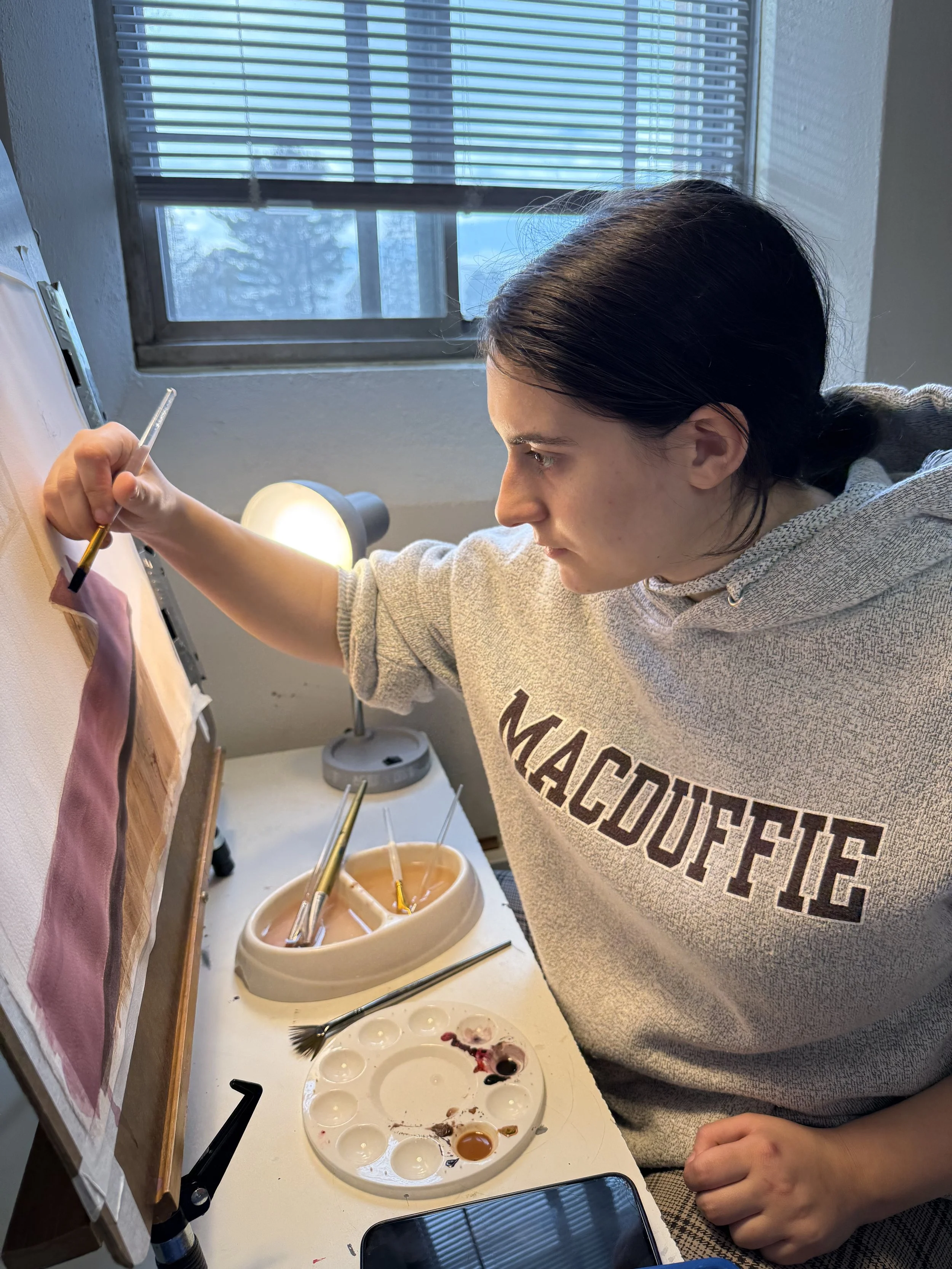 Two women working on pottery at a worktable in a classroom or workshop, with one woman guiding the other in shaping clay.