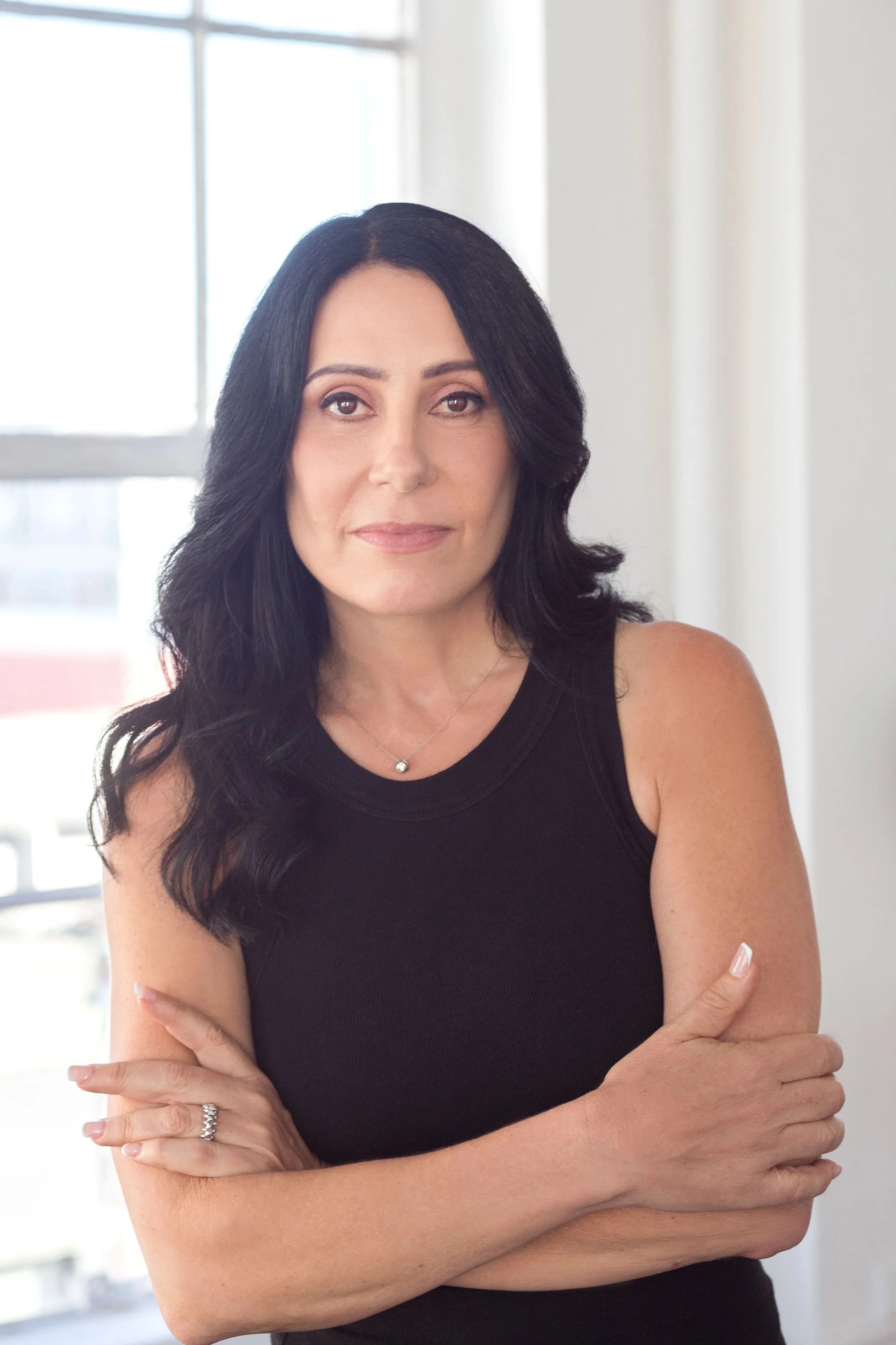 A woman with long black hair wearing a black sleeveless top stands indoors near a window, with her arms crossed and a neutral expression.
