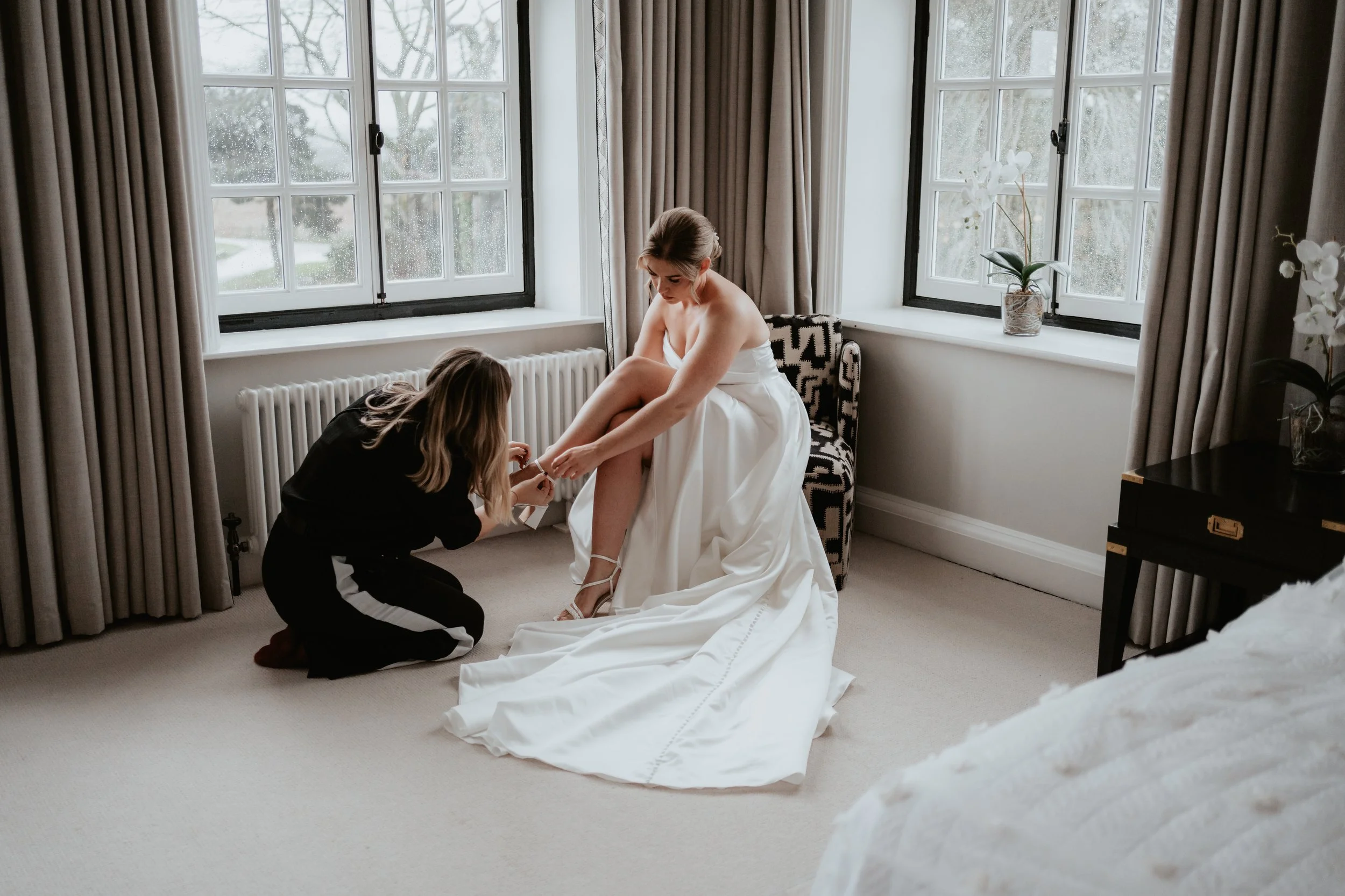 Bride putting on wedding shoes, with wedding coordinator helping