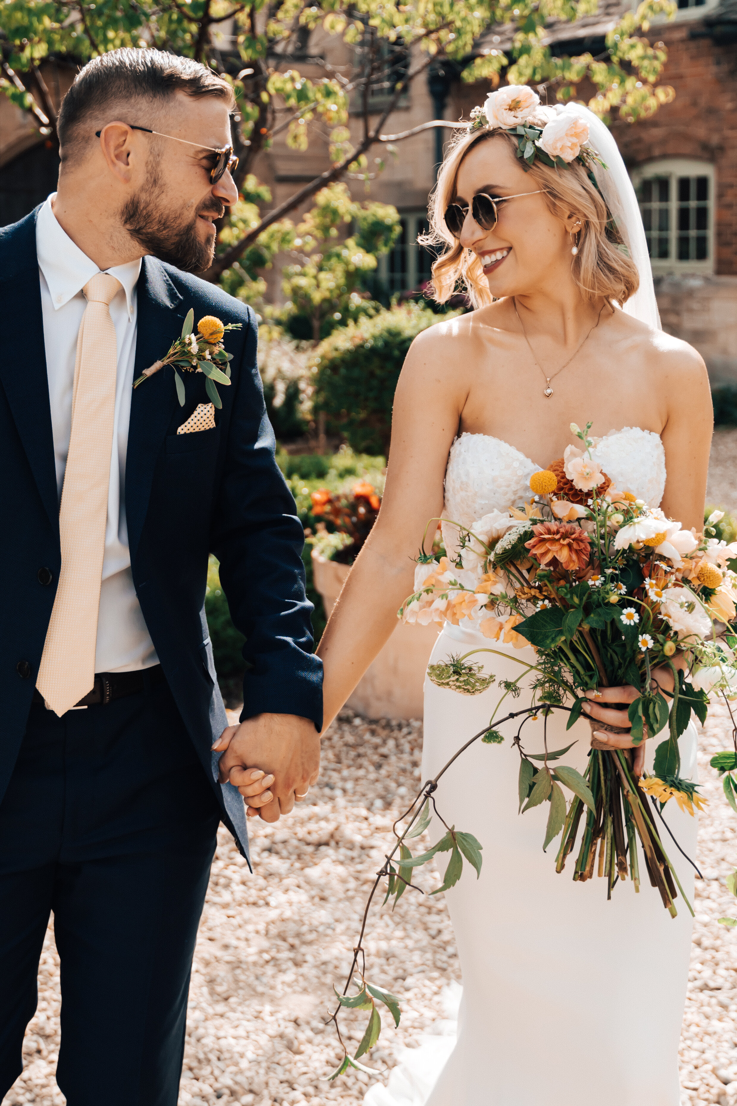A bride and groom holding hands and smiling outdoors on a sunny day, with the bride wearing a strapless white wedding gown, floral crown, and sunglasses, and the groom wearing a dark suit, white shirt, and light-colored tie, with a small boutonniere on his lapel.