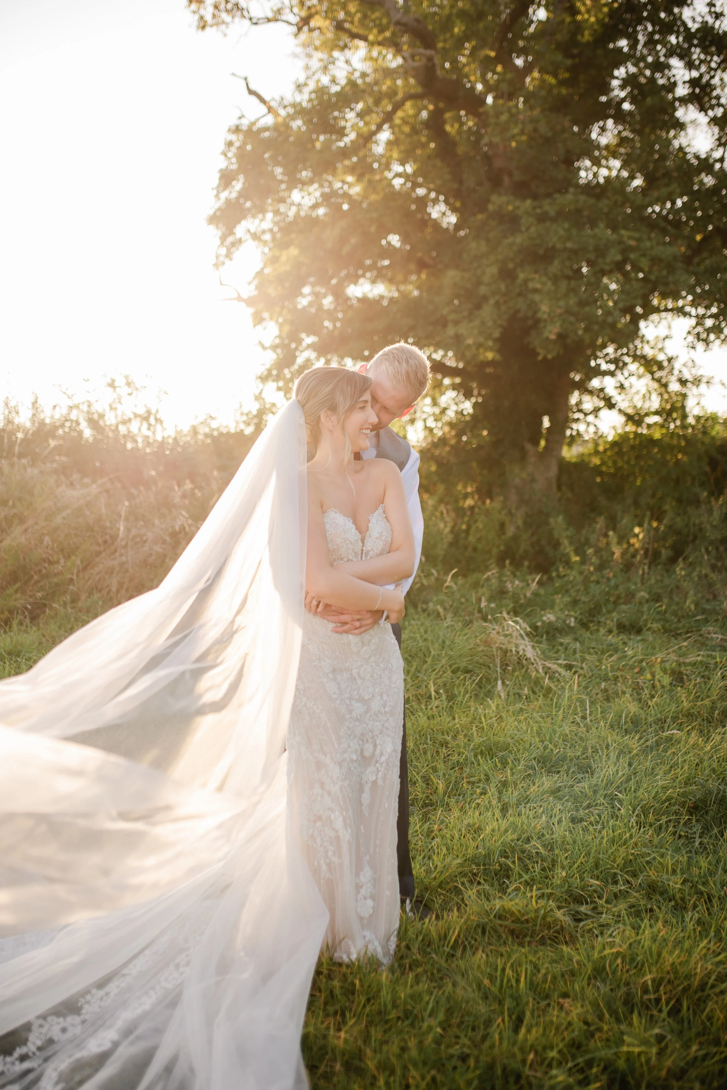 Bride and groom sharing a tender moment outdoors during sunset, with the bride in a lace wedding gown and flowing veil, and the groom in a suit, surrounded by tall grass and a large tree.