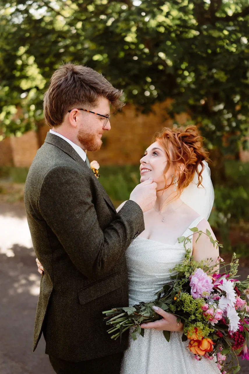 Bride and groom sharing a tender moment outdoors during sunset, with the bride in a lace wedding gown and flowing veil, and the groom in a suit, surrounded by tall grass and a large tree.