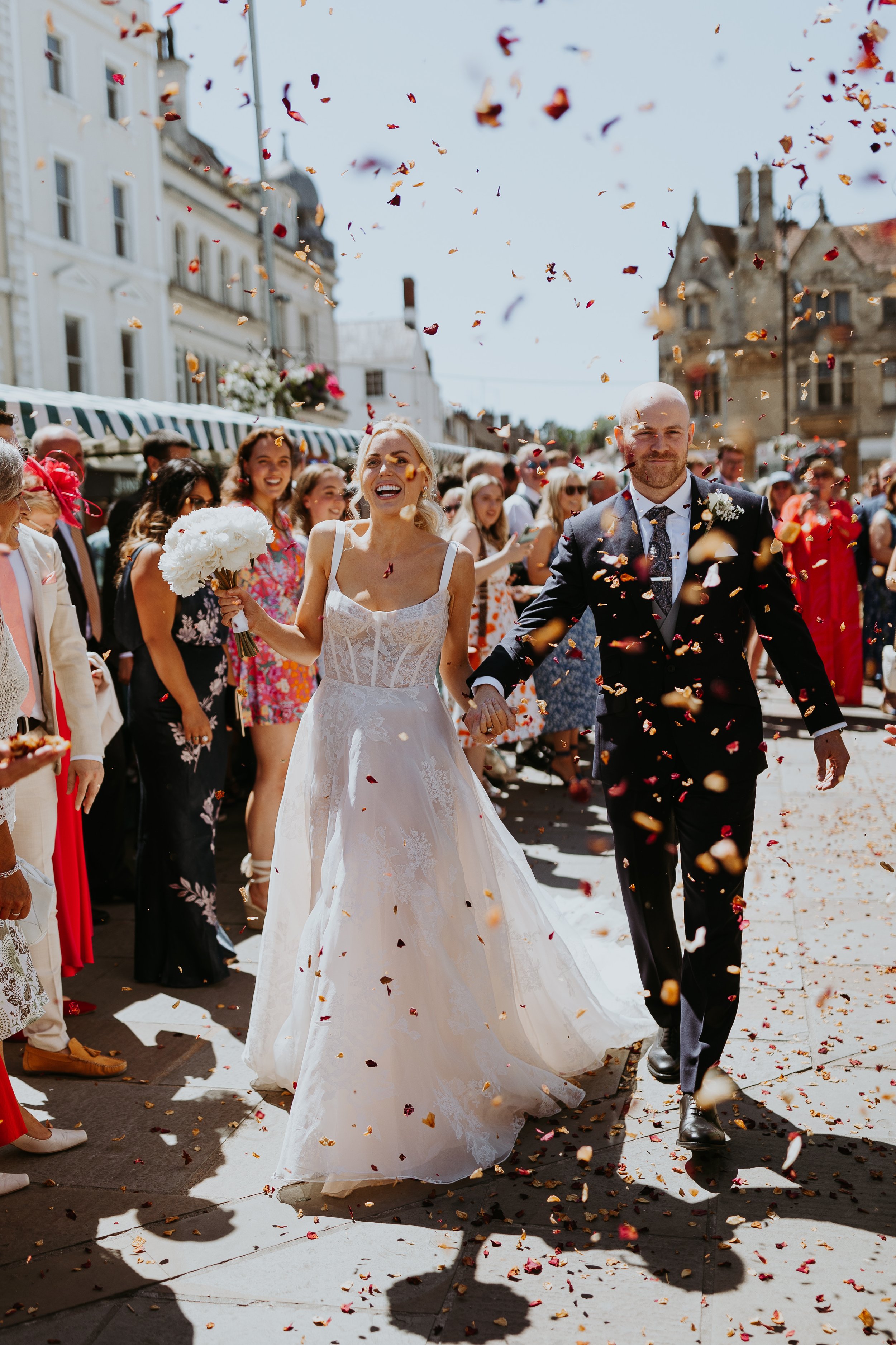 A newlywed couple walking hand in hand through a crowd with falling confetti on a sunny street, celebration with guests in colorful dresses and suits in the background.