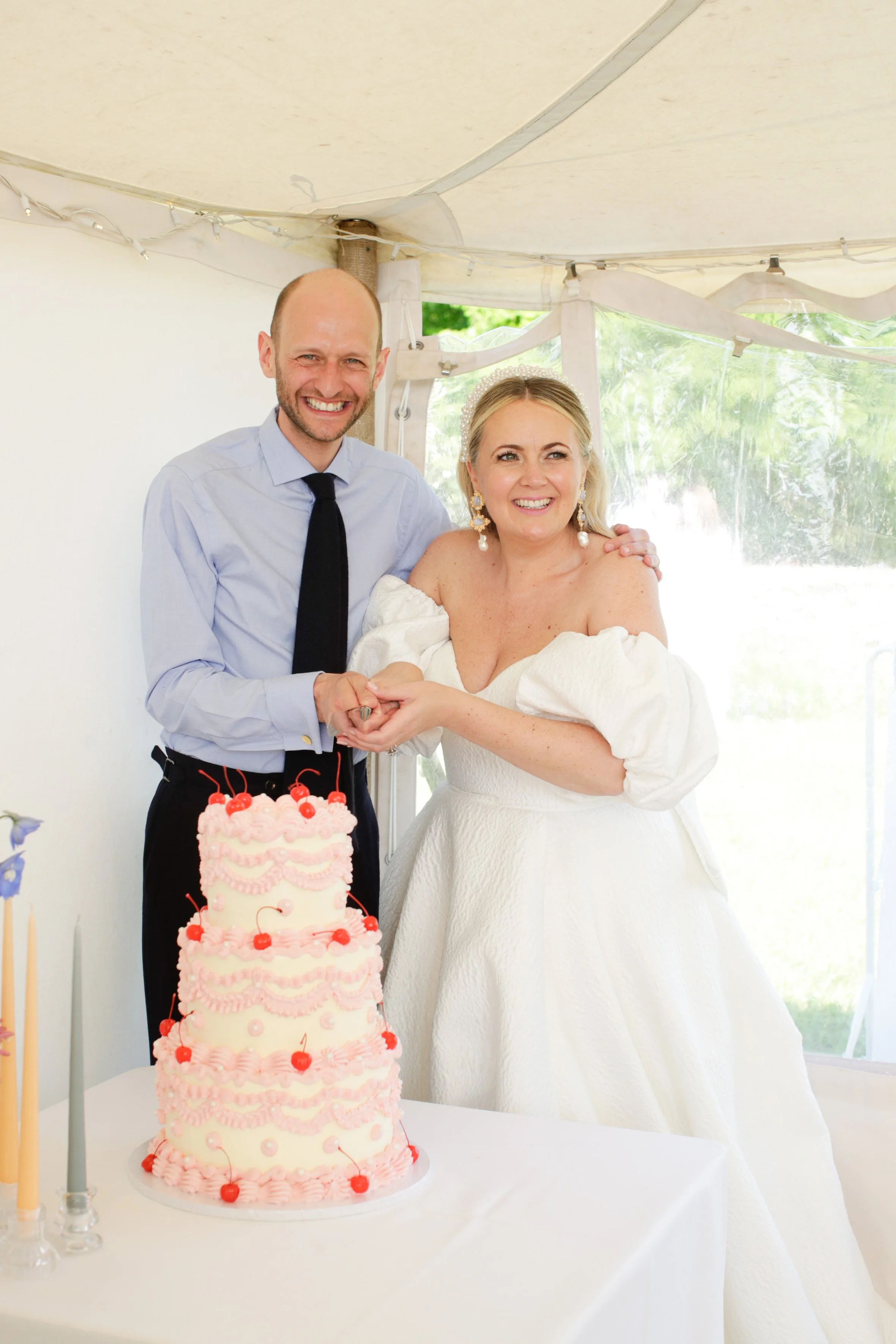 A newlywed couple is smiling and holding hands in front of a four-layer pink and white wedding cake decorated with cherries at their wedding reception.