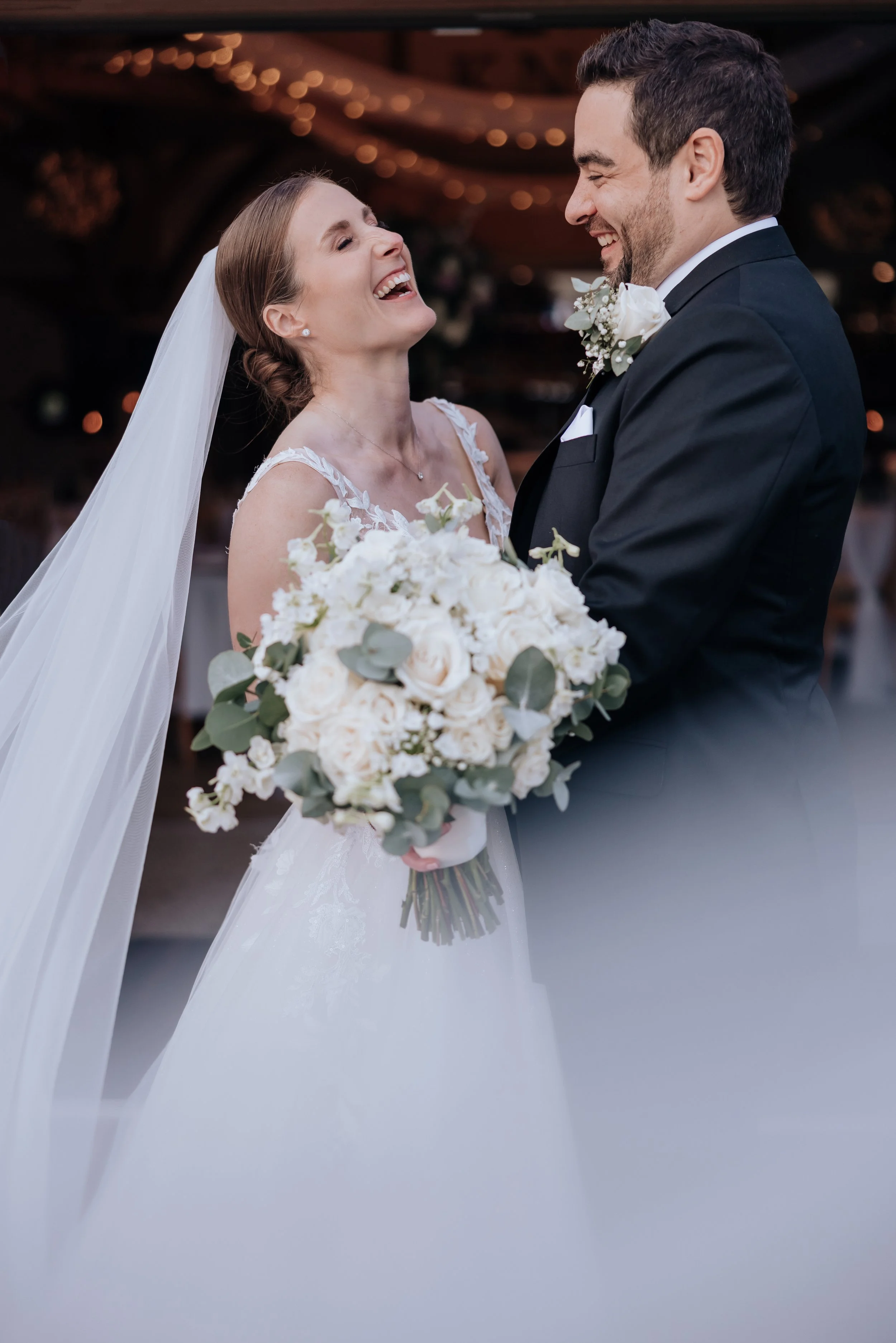 A bride and groom smiling at each other indoors during their wedding, with the bride holding a bouquet of white roses and greenery.