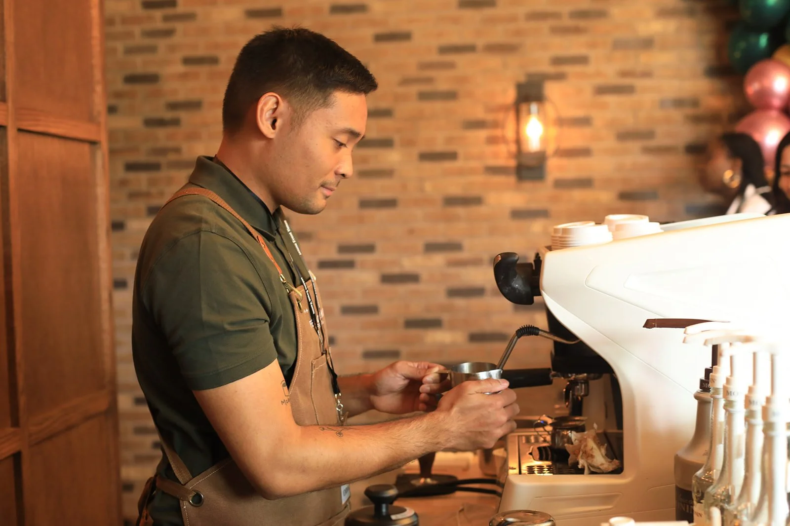 A barista preparing espresso behind a coffee machine in a cafe with a brick wall background and a soft light fixture.