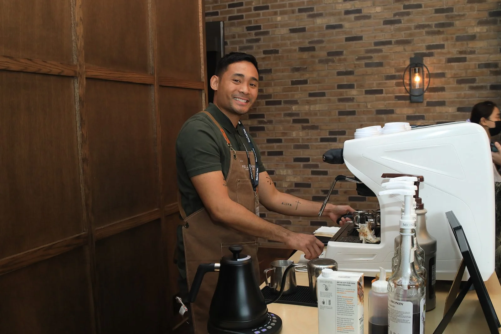 A barista preparing coffee behind a counter at a coffee shop, smiling at the camera, with a brick wall backdrop.