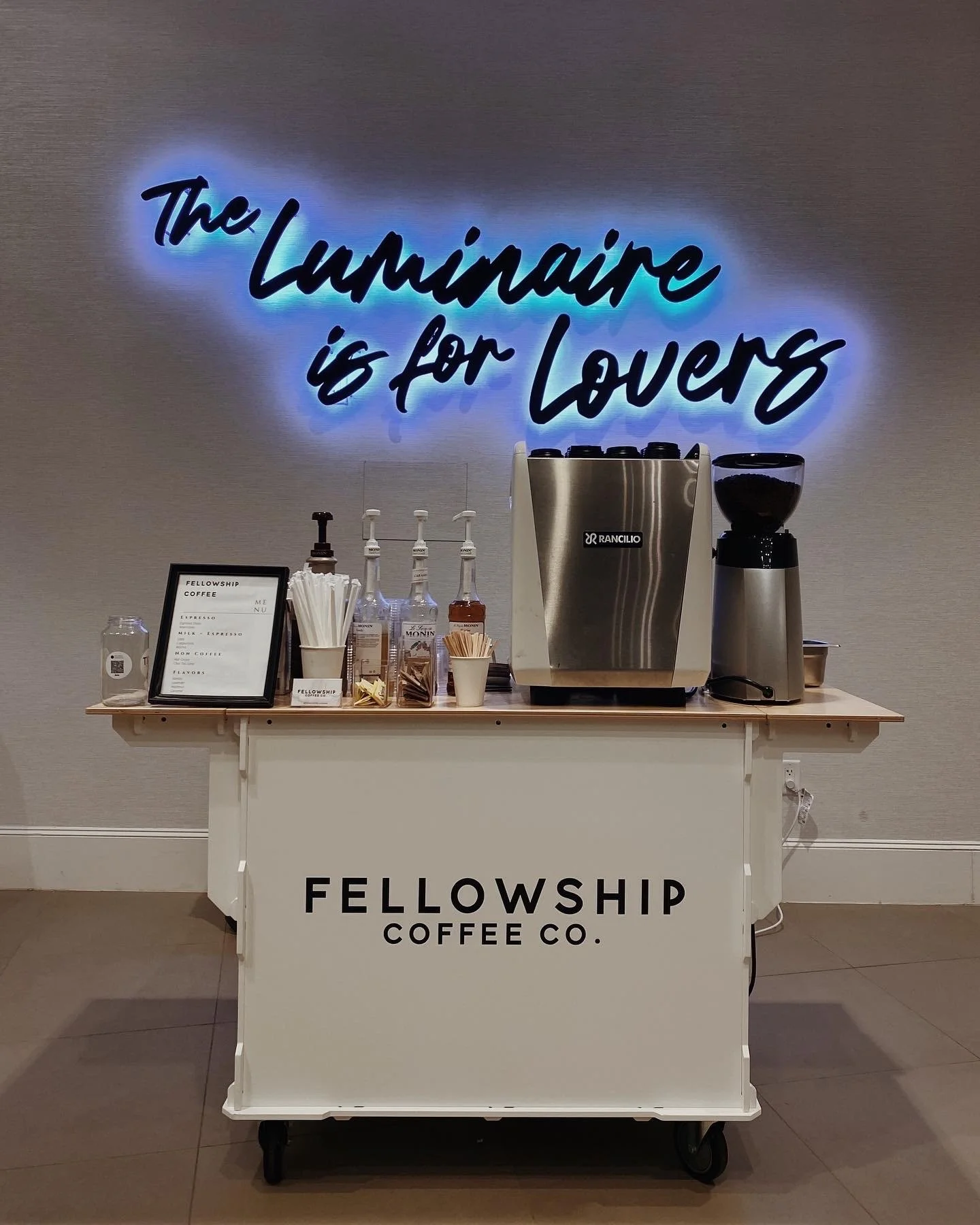 A coffee station with a neon sign that says 'The Luminaire is for Lovers', and a sign that reads 'Fellowship Coffee Co.' on the front of the cart. The station has coffee brewing equipment, bottles of syrup, cups, and stir sticks.