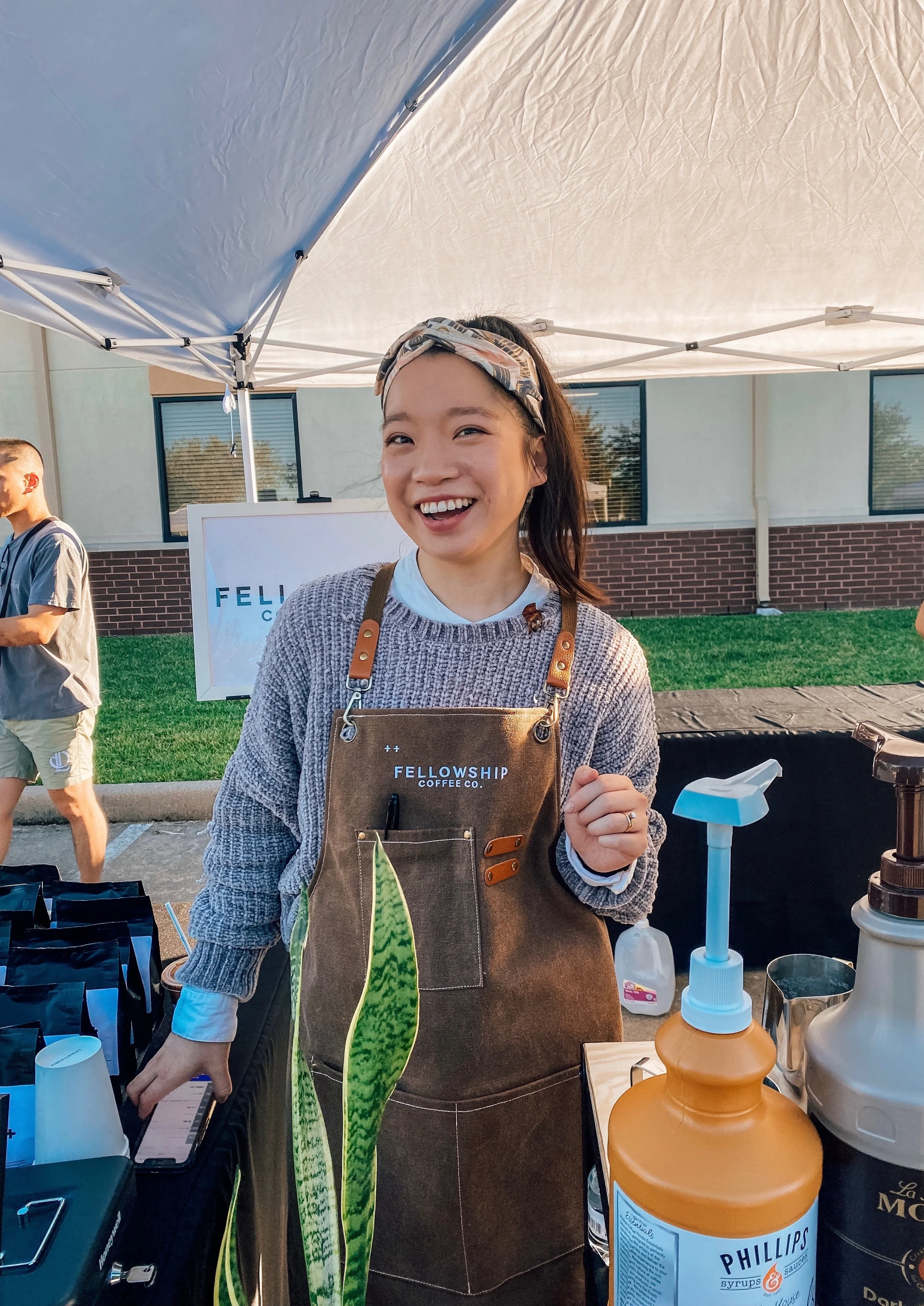 A person smiling while wearing a Fellowship Coffee Co. apron at an outdoor market stall with various beverage supplies under a canopy.