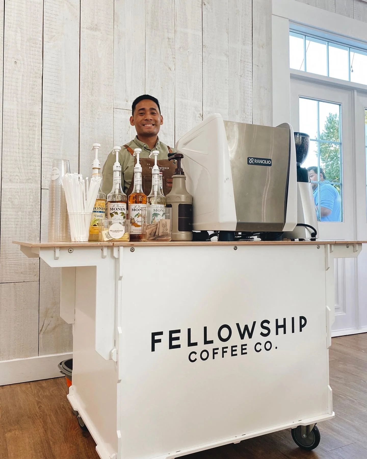 Smiling man standing behind a white mobile coffee cart with the words 'Fellowship Coffee Co.' on it, surrounded by syrup bottles and cups, inside a bright room with wood-paneled wall and large window.