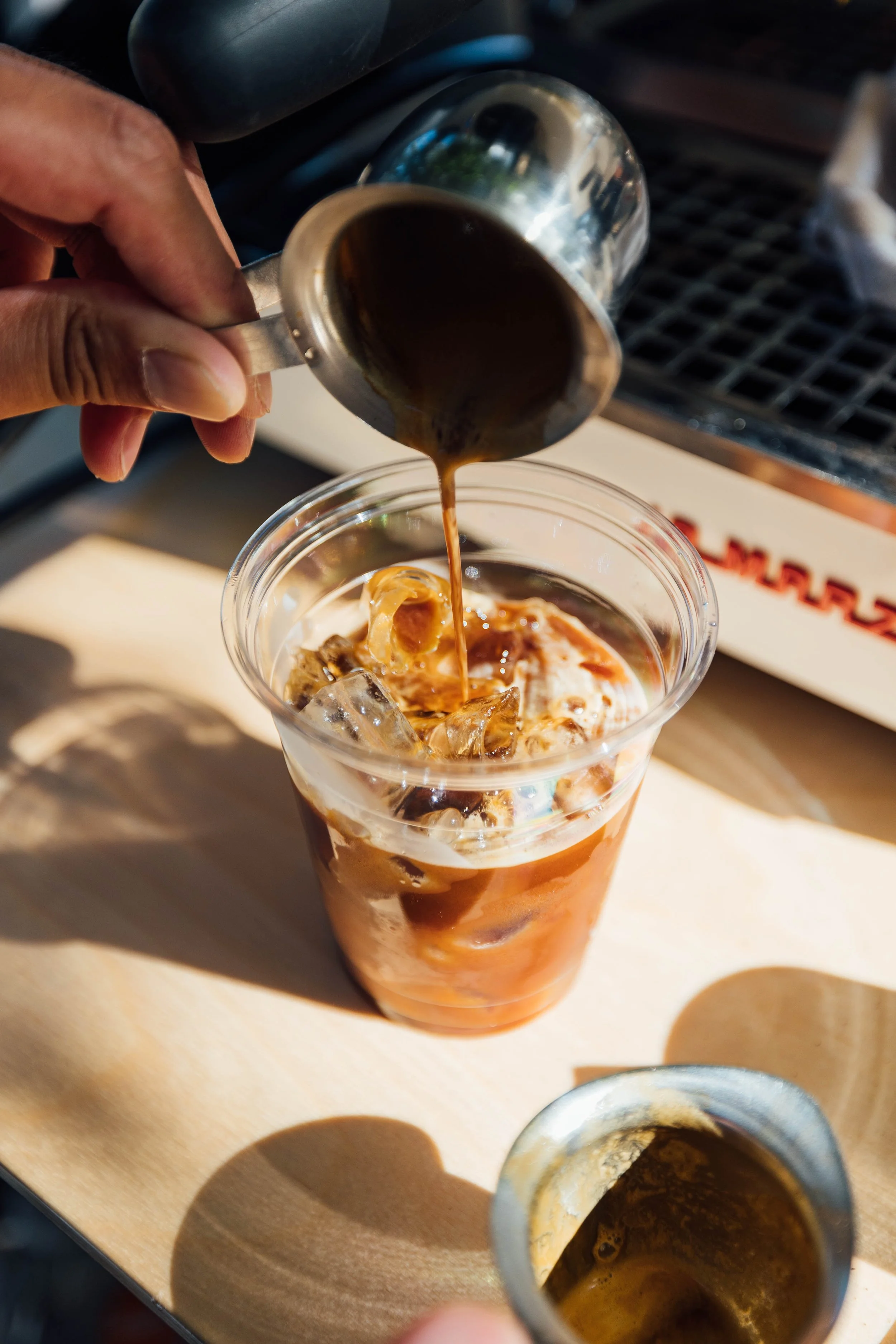 Pouring espresso into a plastic cup with iced coffee and cream on a wooden table.