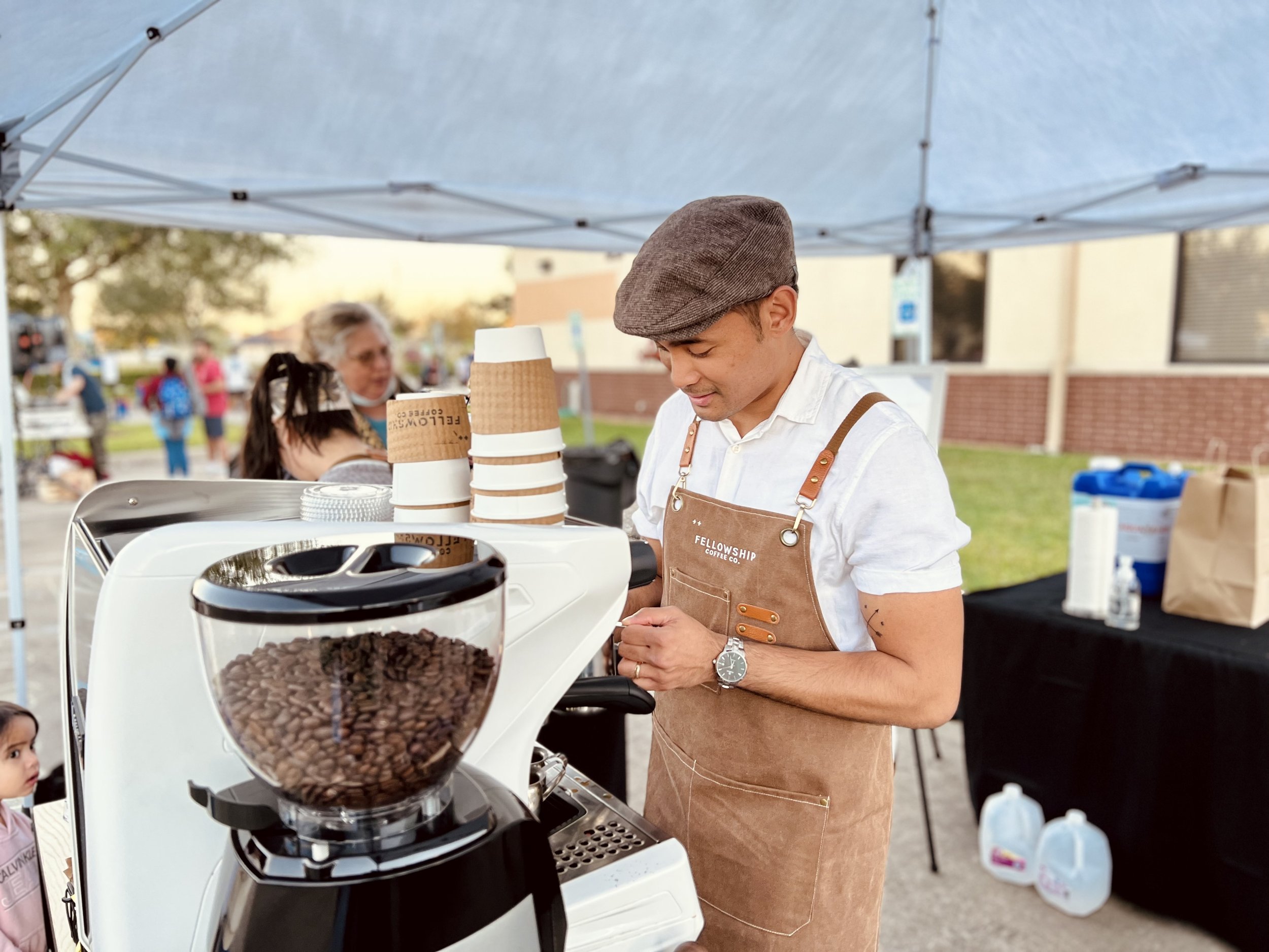 Barista in brown apron making coffee outside under a tent, with a coffee machine and cups.