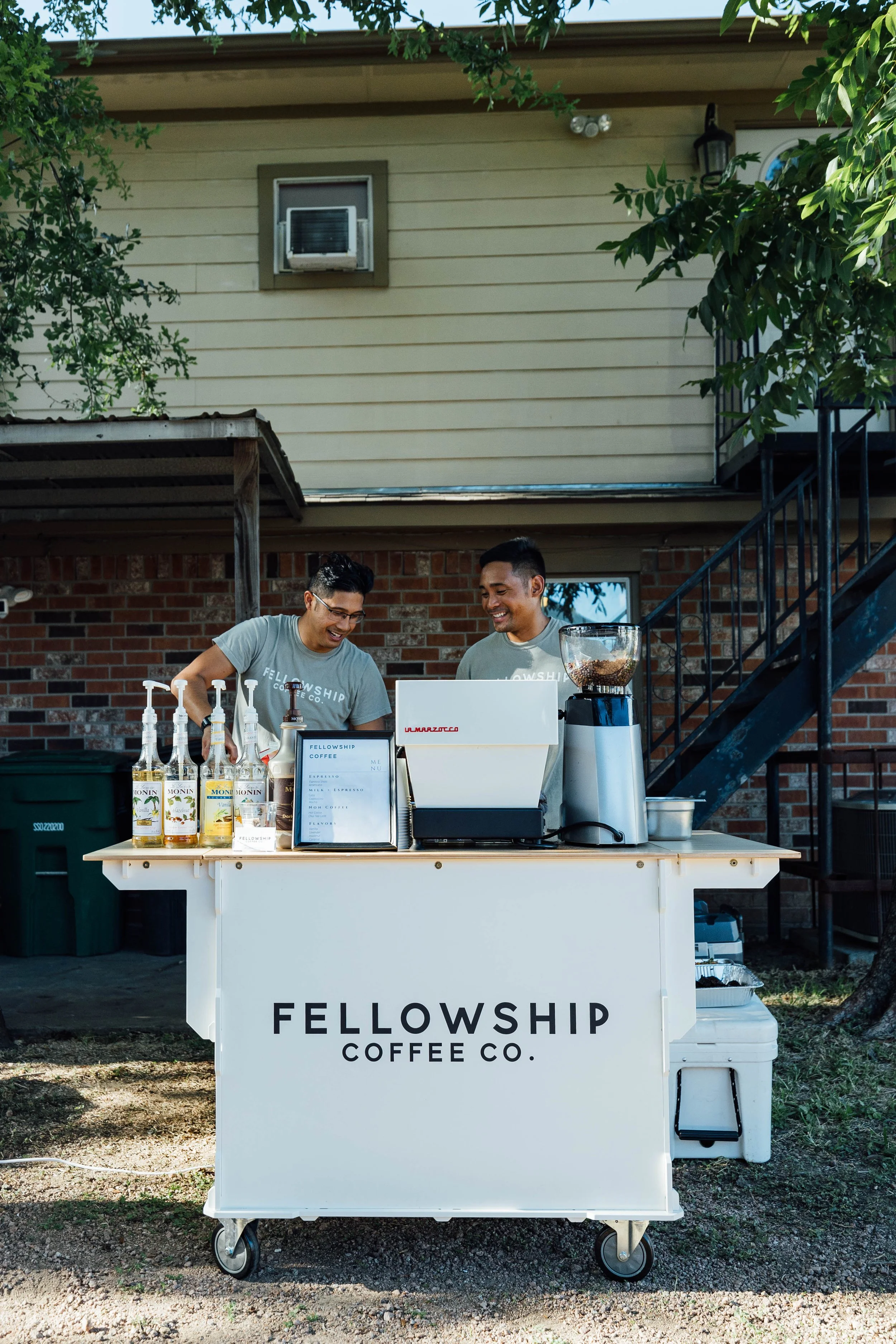 Two men working at a coffee stand with a white table labeled 'Fellowship Coffee Co.' outdoors. The stand has syrup bottles, a menu, and a coffee grinder.