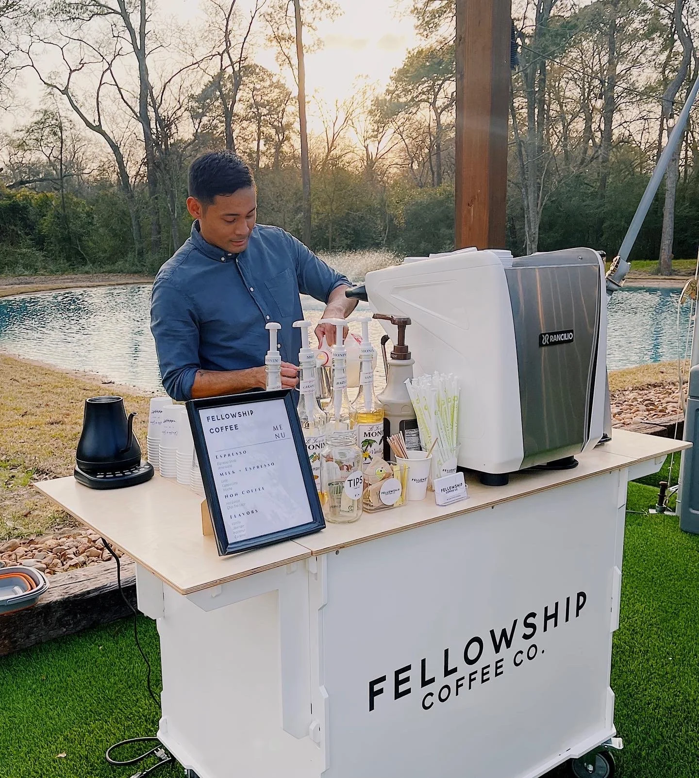 Man preparing coffee at an outdoor station with a lake and trees in the background, labeled Fellowship Coffee Co.
