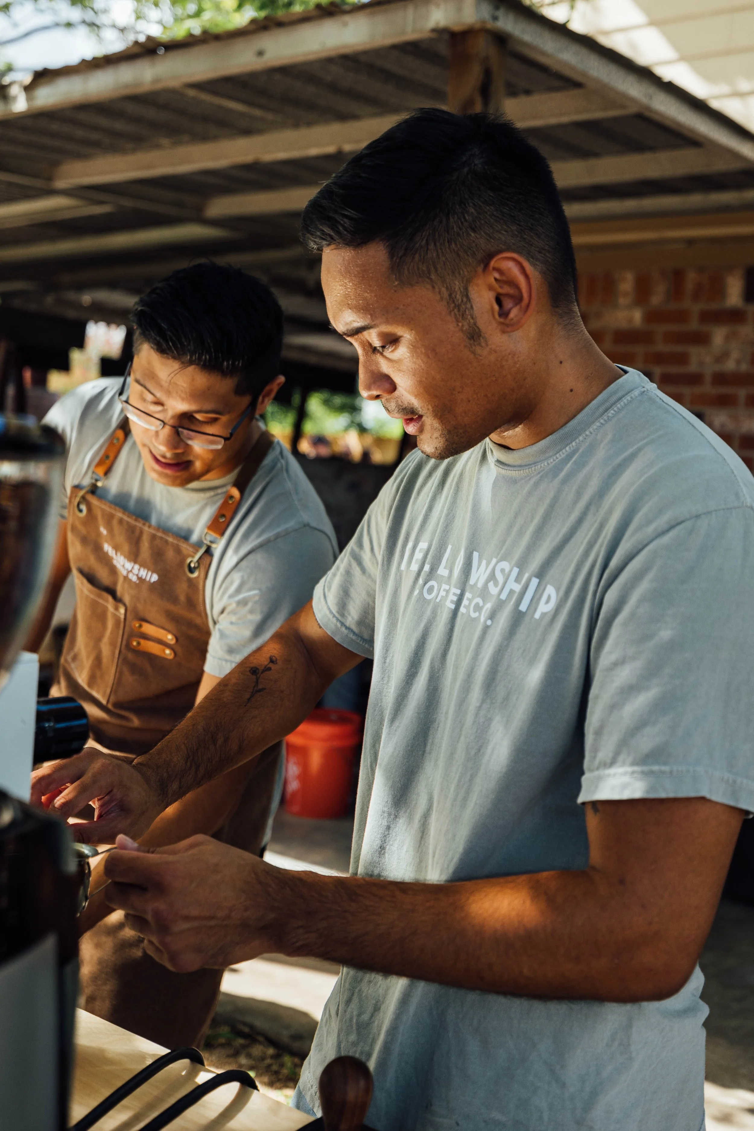 Two men working together in an outdoor kitchen or workshop, one wearing glasses and a brown apron, the other wearing a light gray t-shirt with text, focused on a task, with work tools and equipment around them.