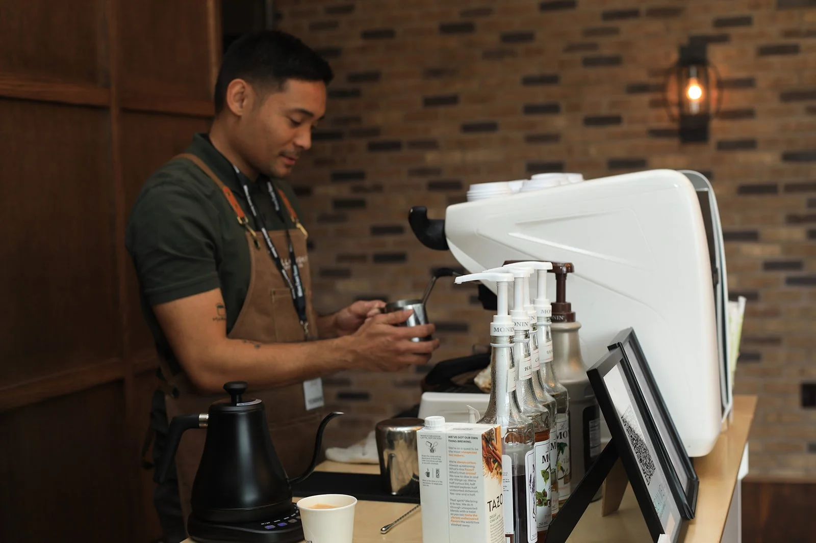 Barista preparing coffee behind a counter with coffee bottles, a kettle, and a digital menu display.