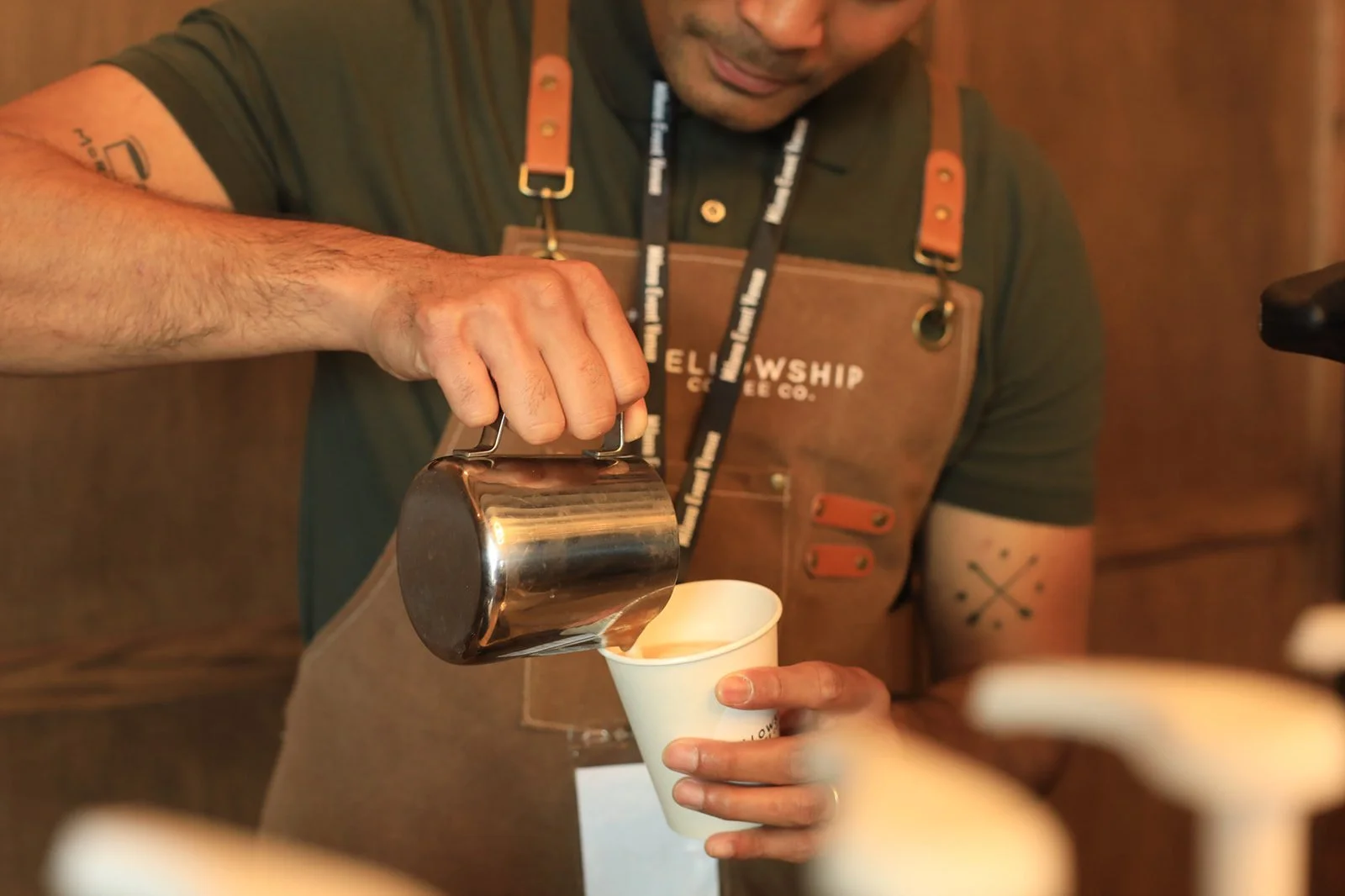A barista pouring hot liquid from a stainless steel pitcher into a white paper cup.