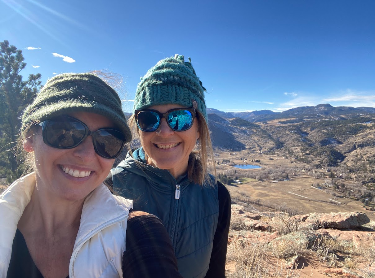 Two smiling women wearing sunglasses and winter hats take a selfie on a mountain with a landscape of mountains, a lake, and a valley in the background under a blue sky.