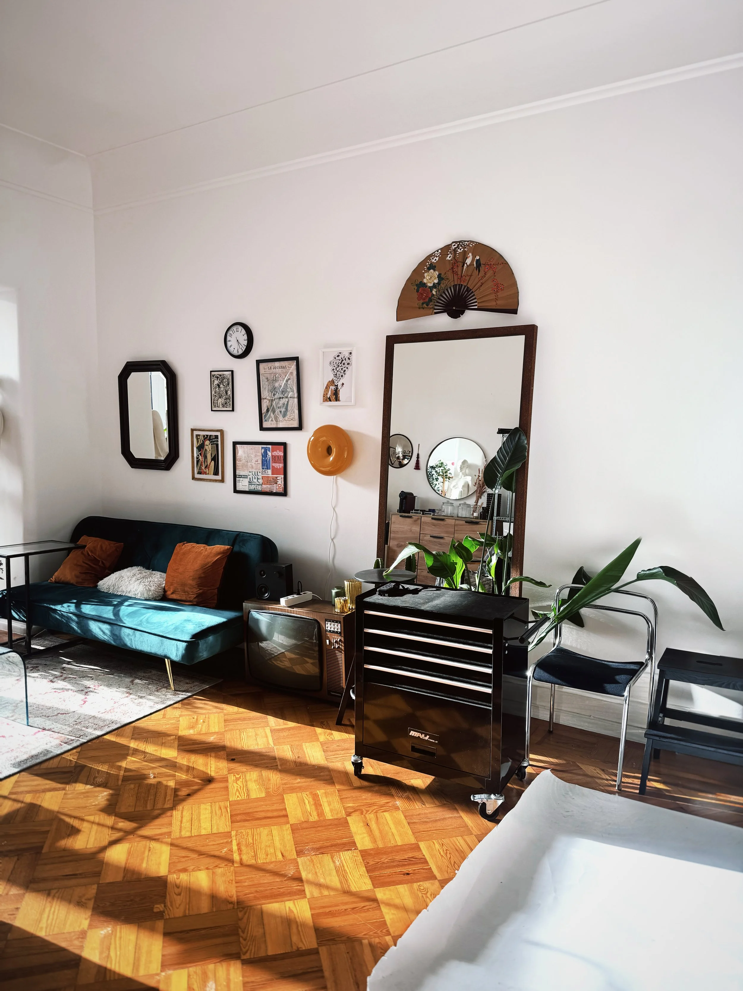 Living room with a blue velvet sofa, orange pillows, wall art, large mirror, wooden furniture, plants, and sunlight on hardwood floor.