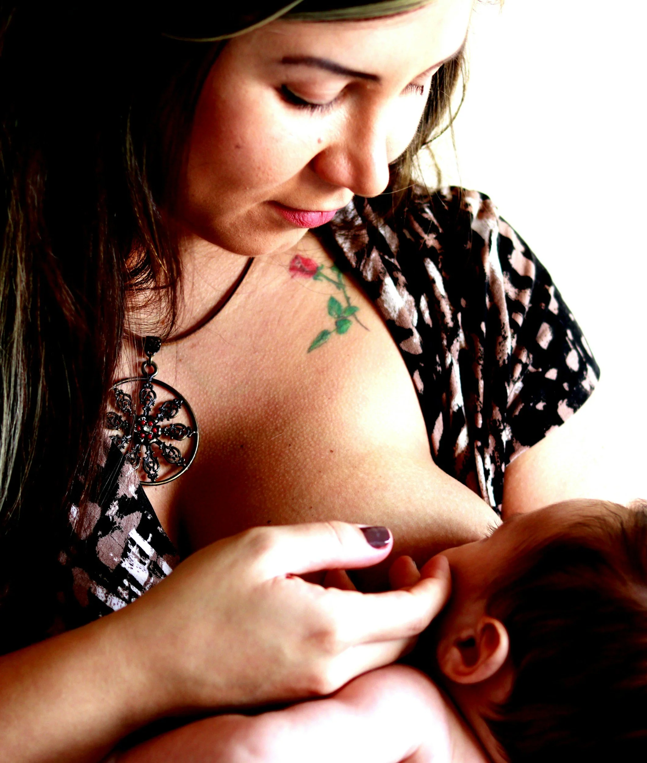 A woman with dark hair and a floral tattoo on her shoulder breastfeeds a baby, wearing a patterned top and jewelry.