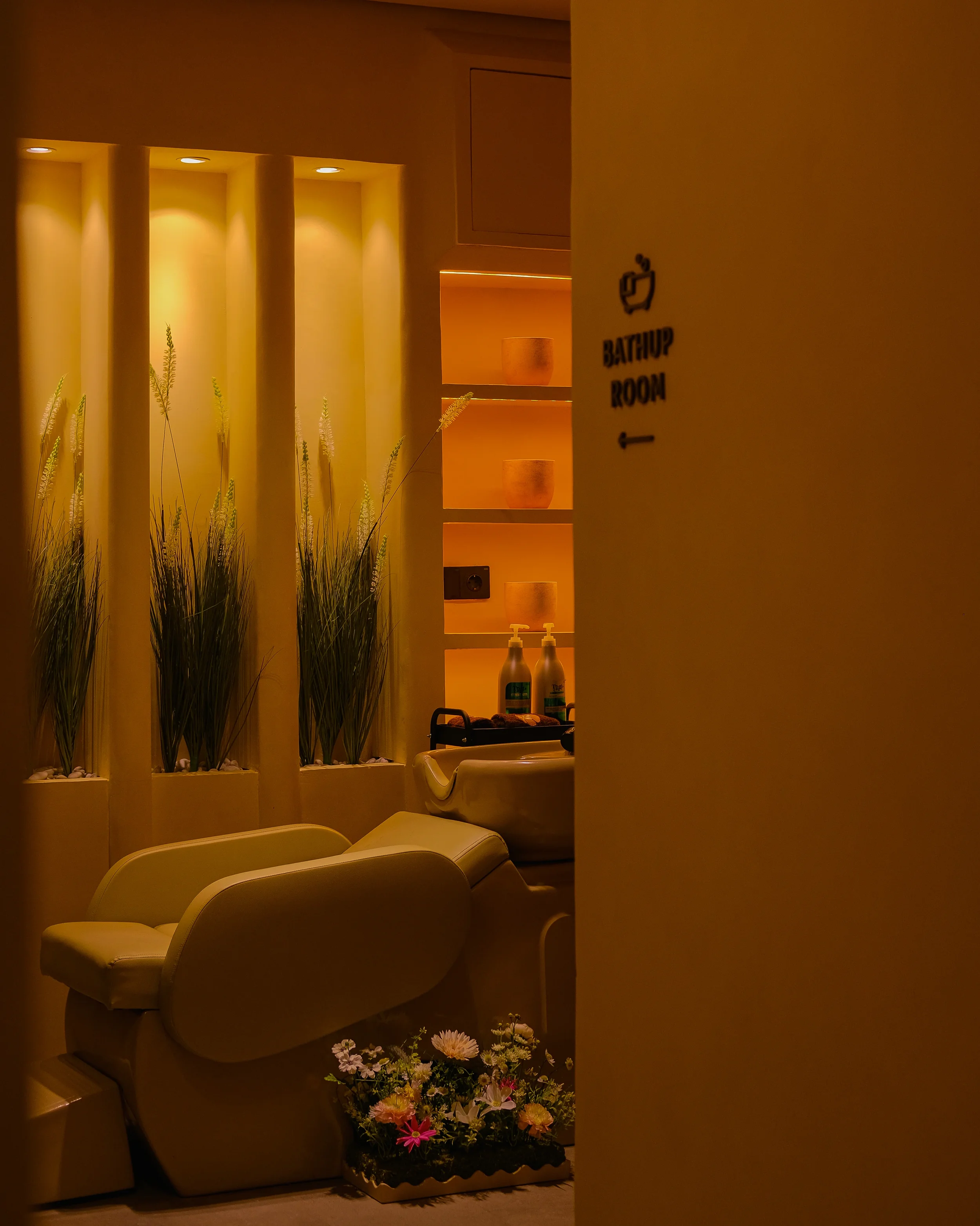 A dimly lit room with beige chairs, green plants, and shelves with decorative vases. A sign indicates the bathroom is to the left.