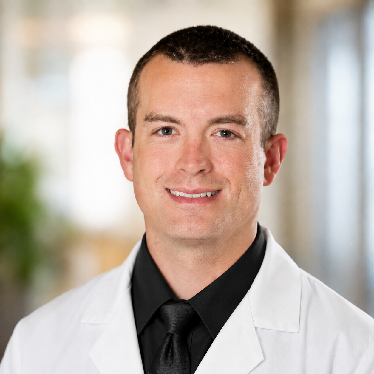 A young man with short dark hair, wearing a white medical coat and black shirt with a black tie, smiling in a professional setting.