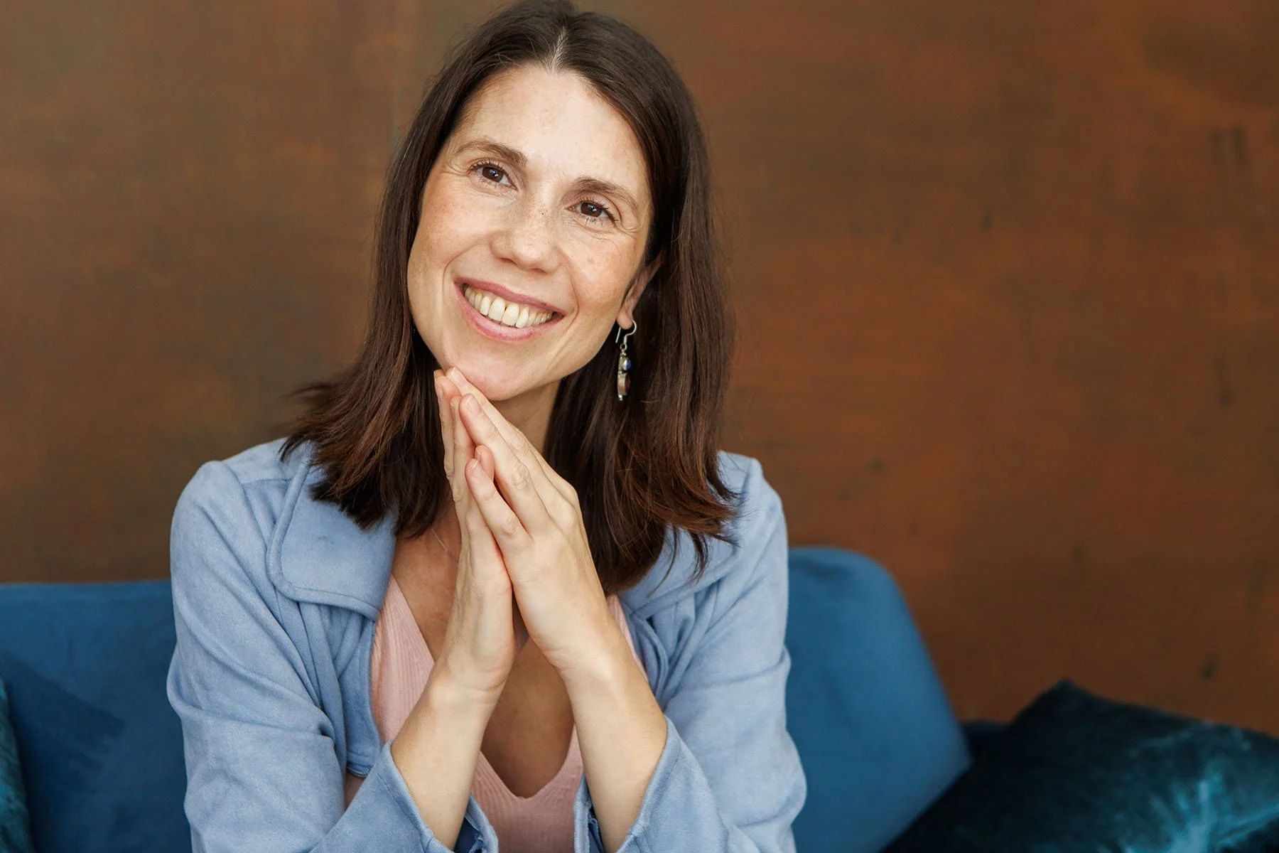 Verónica, sonriente con las manos juntas, sentada en un sofá, con fondo de pared de color marrón y cojines azules. Veronica Civatti, Terapia Gestalt, Eneagrama, Constelaciones Familiares y Somatic Experiencing en Barcelona
