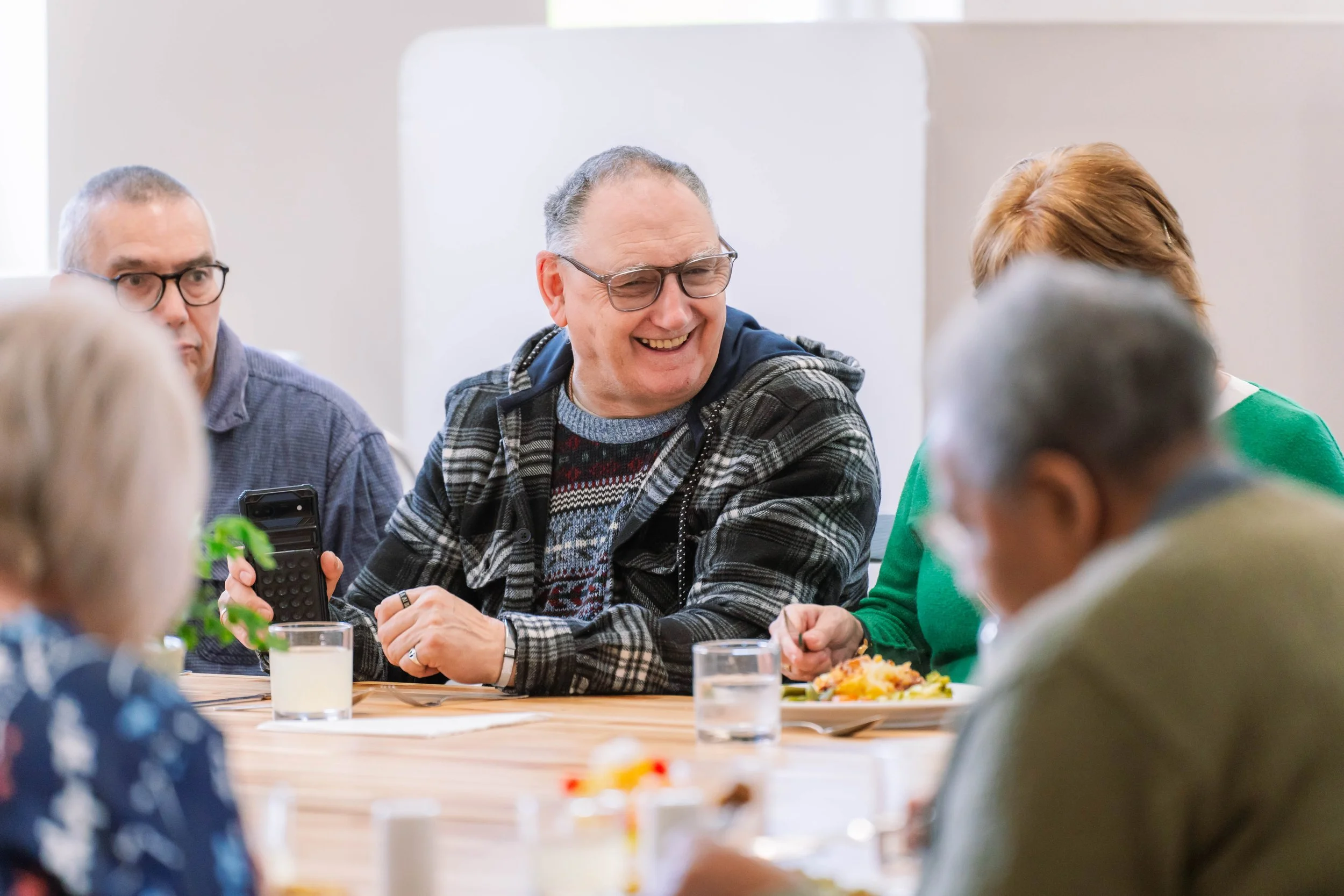 A group of middle-aged to elderly people sitting at a dining table, engaged in conversation and smiling, with some holding utensils and glasses of drinks, in a well-lit room.