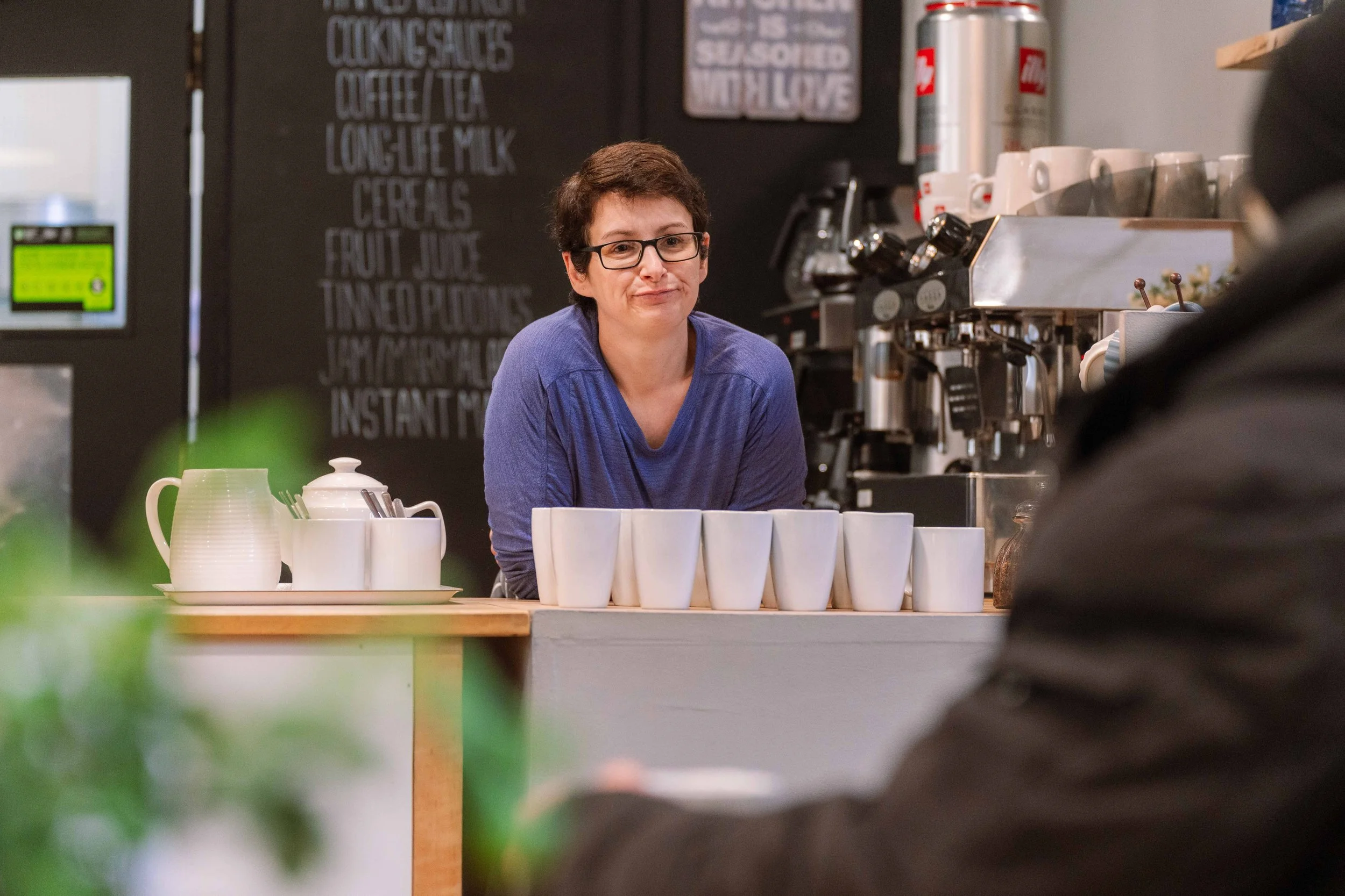 Barista with short dark hair, glasses, wearing a blue shirt, standing behind a coffee counter with white cups, a teapot, and a milk pitcher, in a coffee shop.