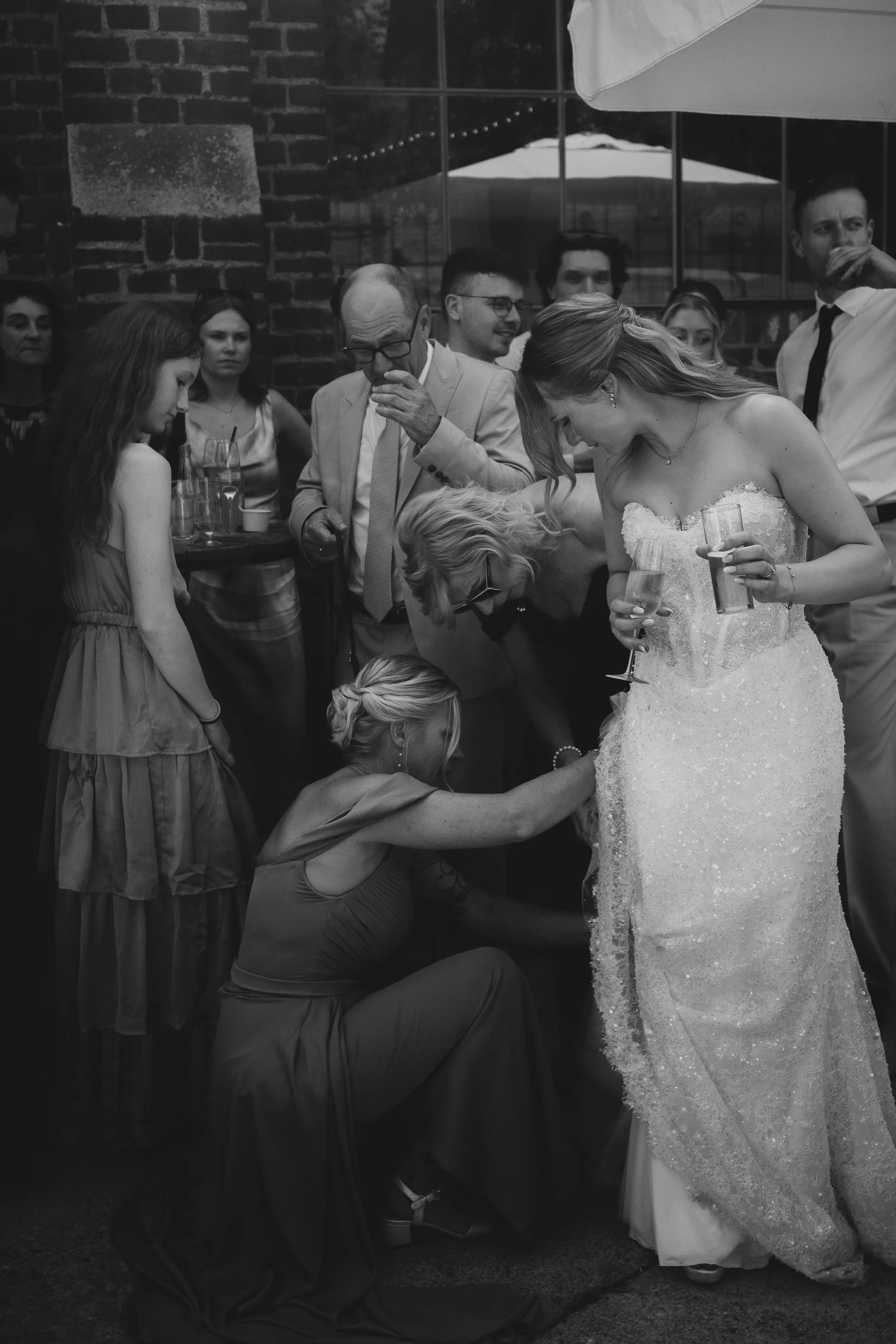 Eine Hochzeitsfeier im Innenraum mit mehreren Gästen, die die Braut und den Bräutigam umringen. Die Braut trägt ein elegantes, trägerloses Brautkleid und hält ein Glas Champagner, während eine Person vor ihr auf den Knien anscheinend an ihrer Schuhe arbeitet.