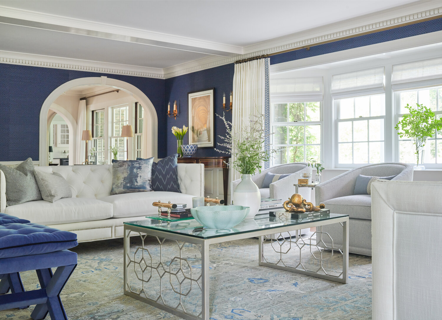 Bright blue living room with white sofa, glass coffee table, patterned rug, and arched doorway, showcasing a refined interior design by Georgia Zikas, featured in Mansion Global.